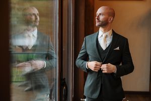 Groom in dark suit adjusts his cuffs while standing near window with reflection.