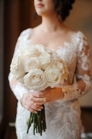 A close-up view of a white wedding bouquet with roses being held against a lace wedding dress.