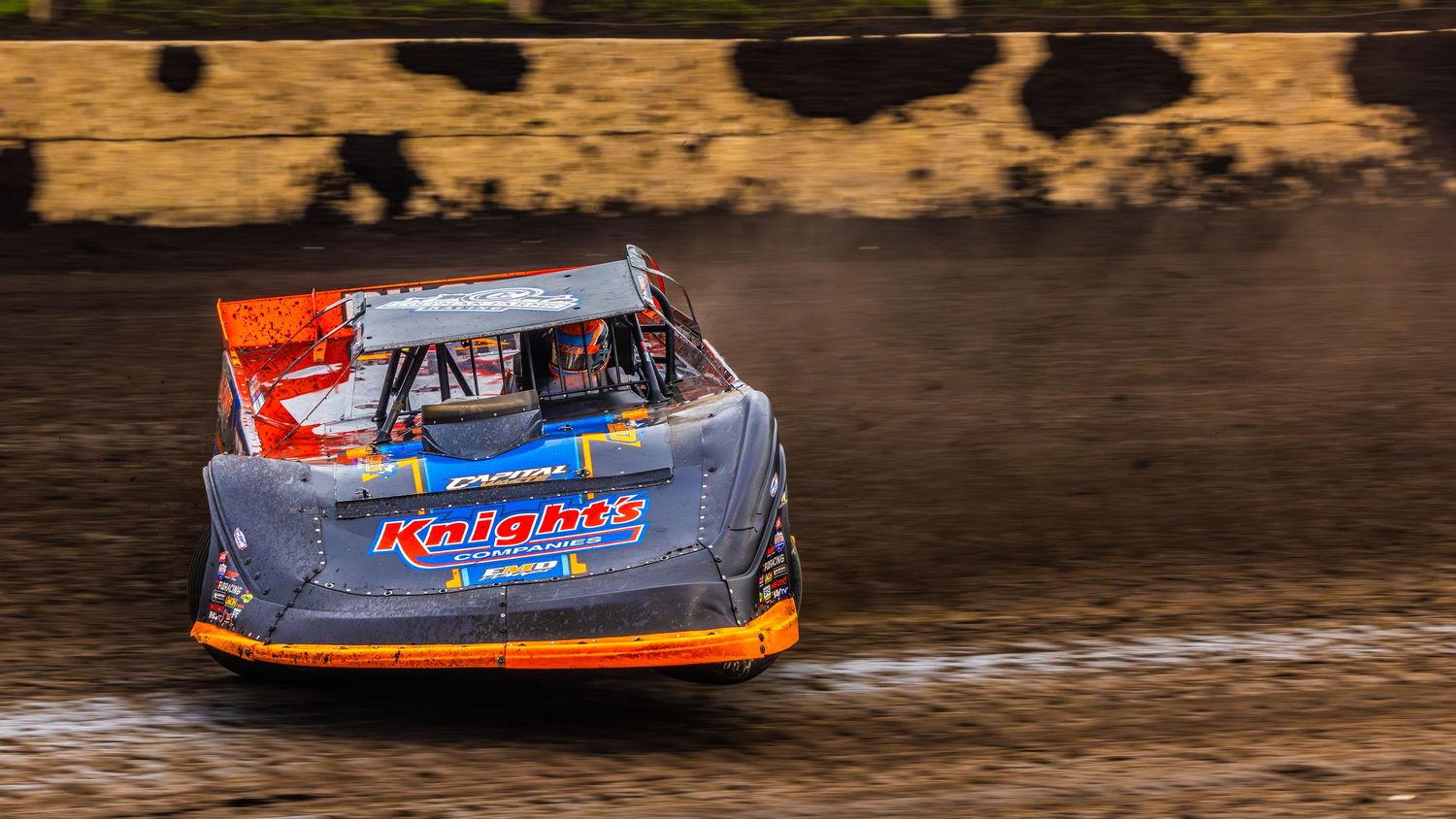 Racing fans watch from crowded grandstands as a Huber's sponsored race car speeds past at a dirt track speedway event.
