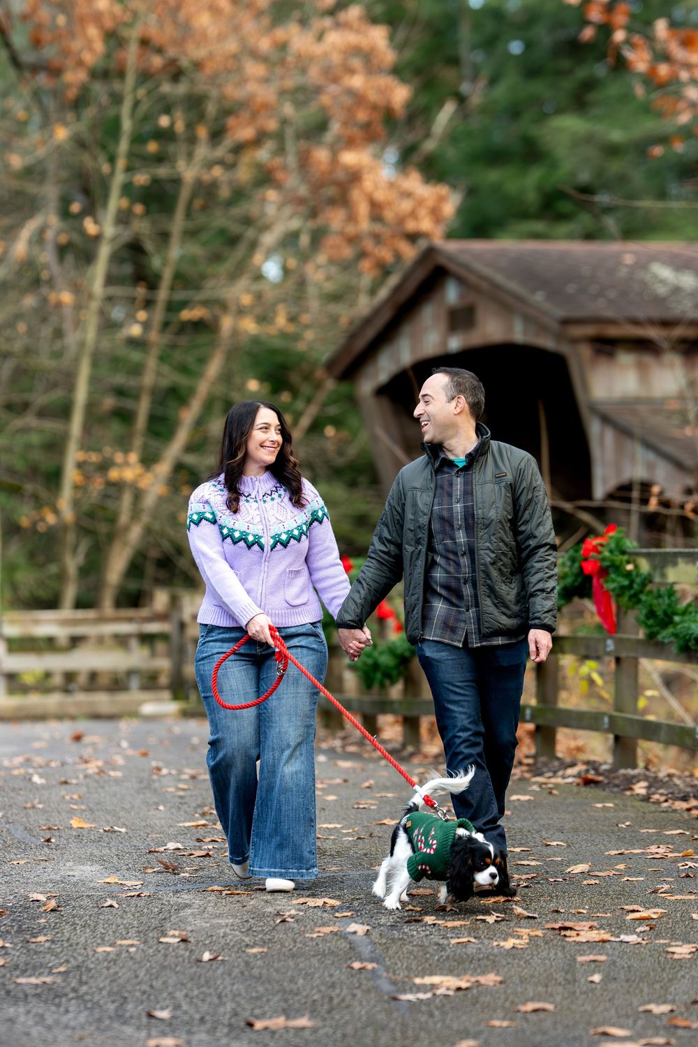 Smiling couple walking their Cavalier King Charles Spaniel on a leash near a covered bridge with holiday decor.