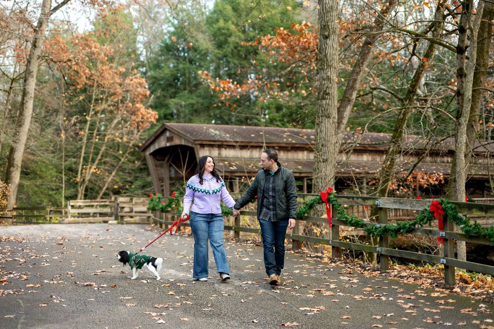 Couple walking their small dog on a leaf-covered path near a festively decorated covered bridge in autumn.