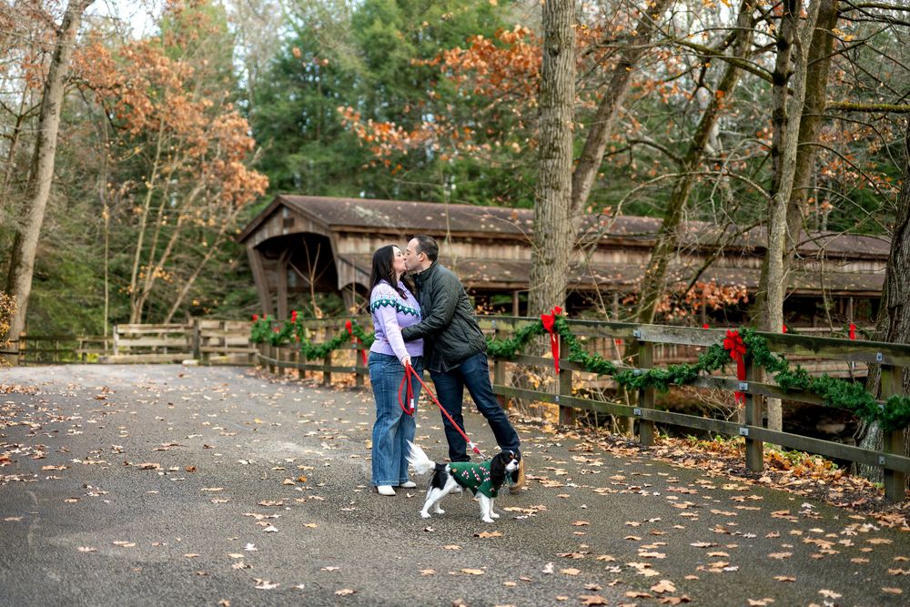 Couple sharing a kiss while walking their dog on a scenic autumn path near a Christmas-decorated covered bridge.