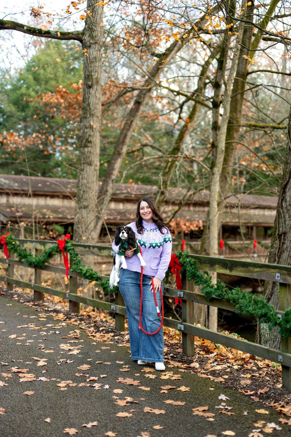 Smiling woman holding a Cavalier King Charles Spaniel on a leash near a Christmas wreath-adorned wooden fence.