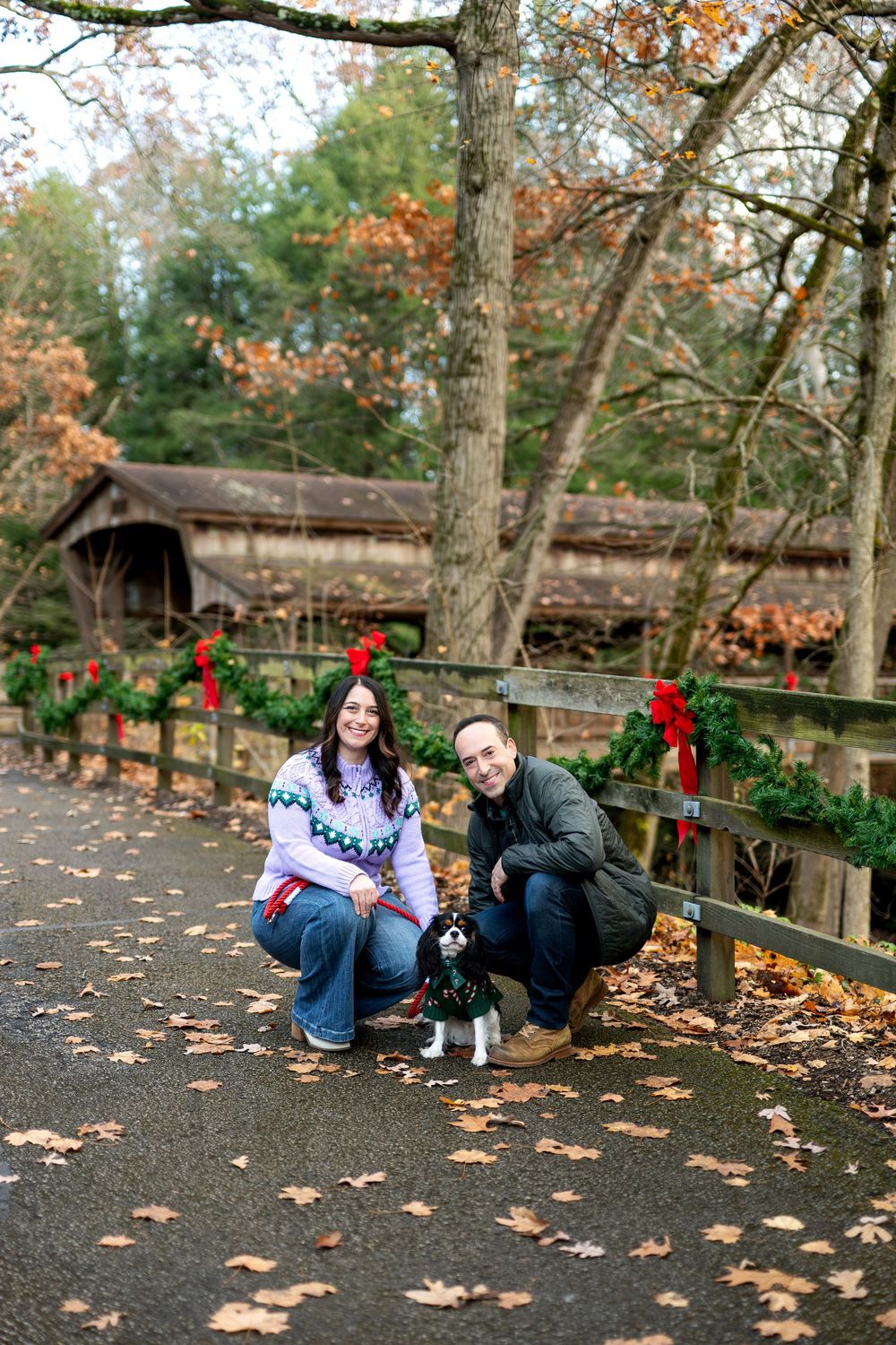 Couple kneeling with their Cavalier King Charles Spaniel near a Christmas wreath-lined fence and covered bridge.