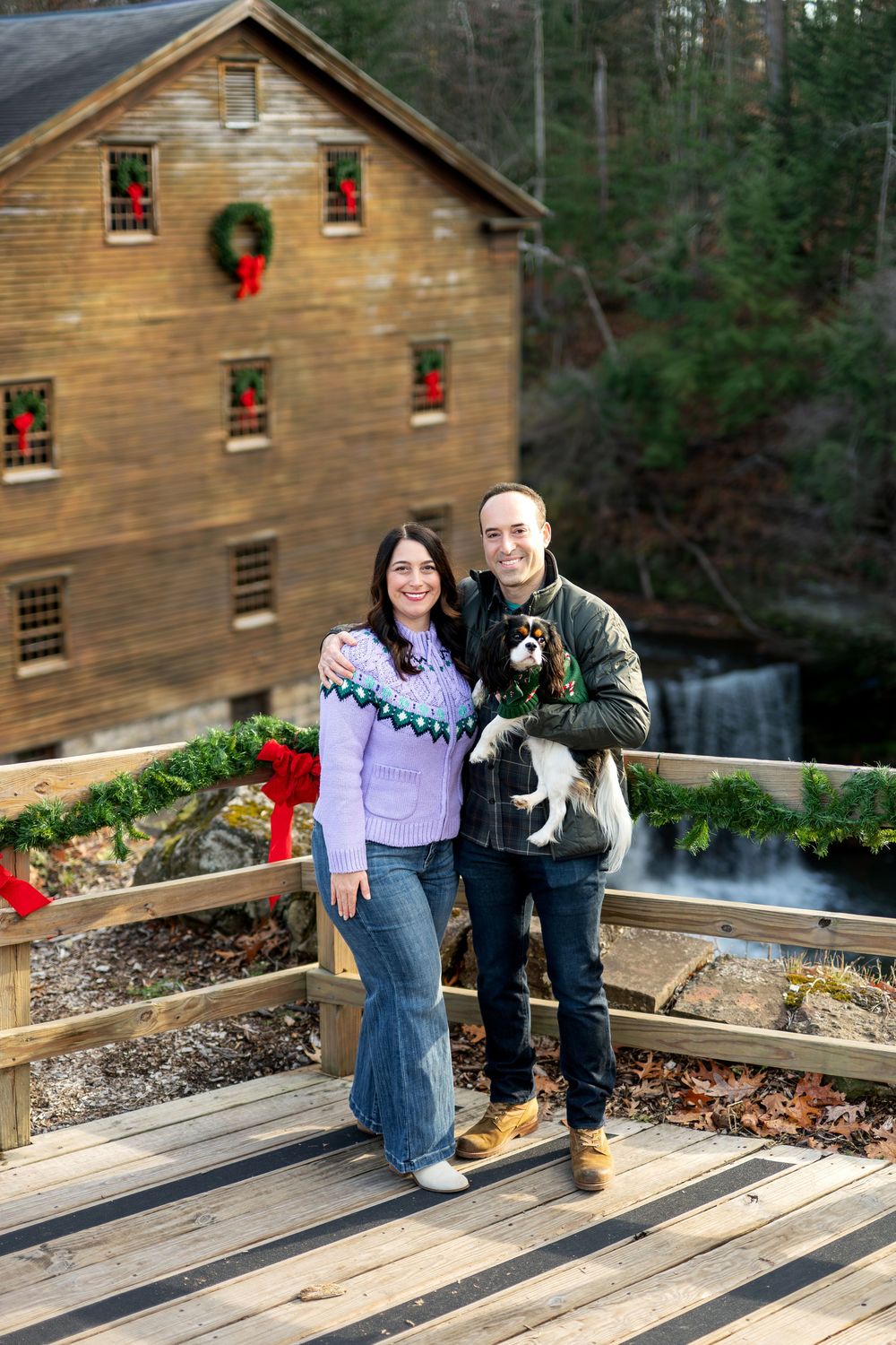Smiling couple holding Cavalier King Charles Spaniel at Christmas-decorated historic mill bridge.