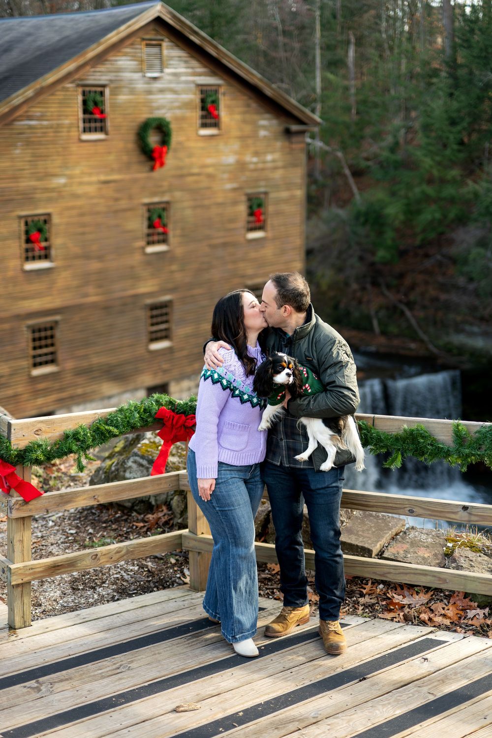 Couple sharing kiss while holding dog on festive holiday bridge with mill and waterfall.