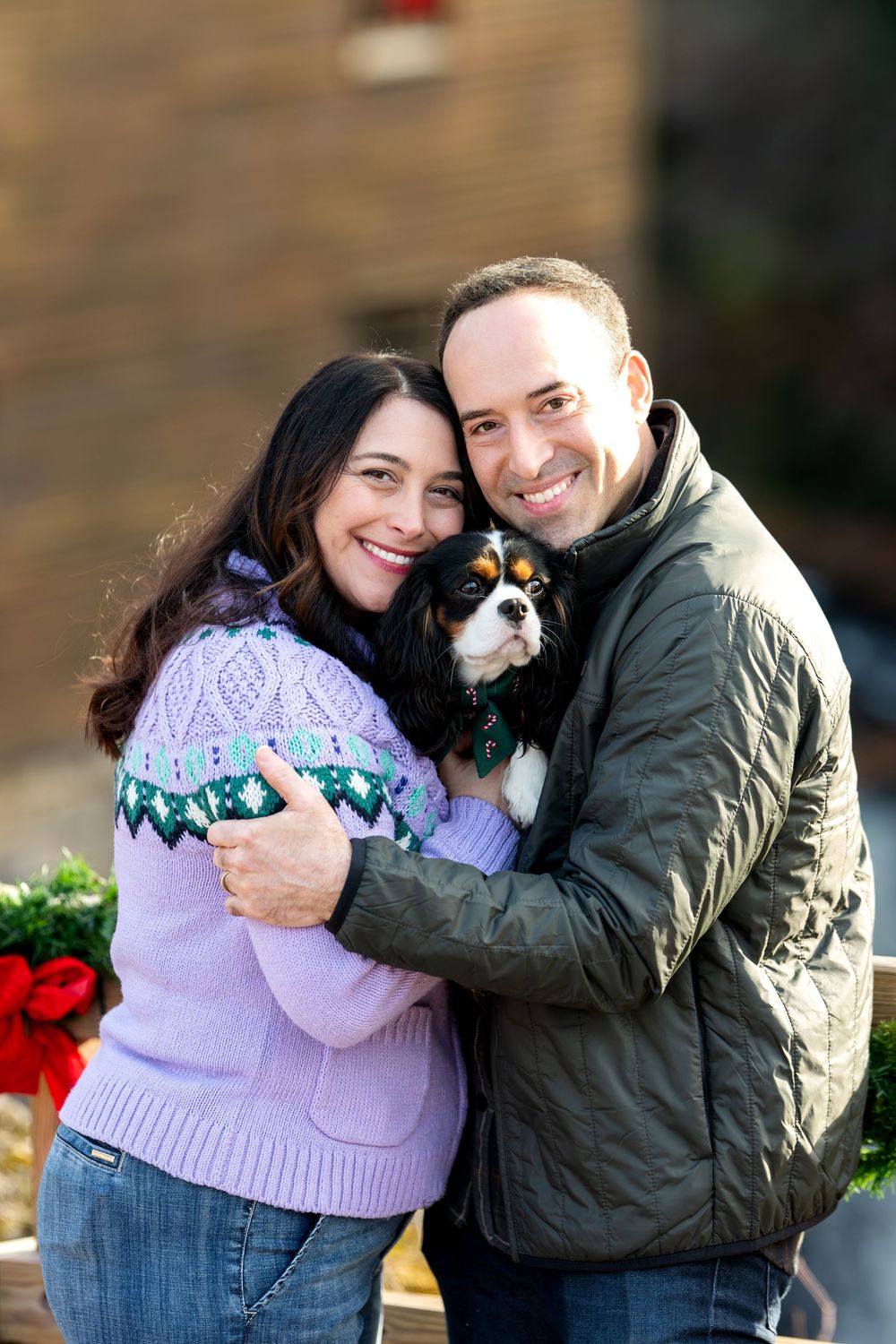 Cheerful couple cuddling Cavalier King Charles Spaniel with Christmas decorations in background.