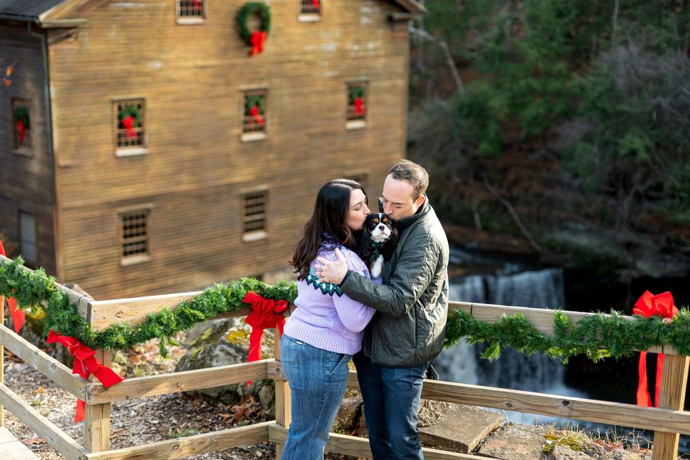 Happy couple dancing together on Christmas-decorated bridge near historic stone grist mill.
