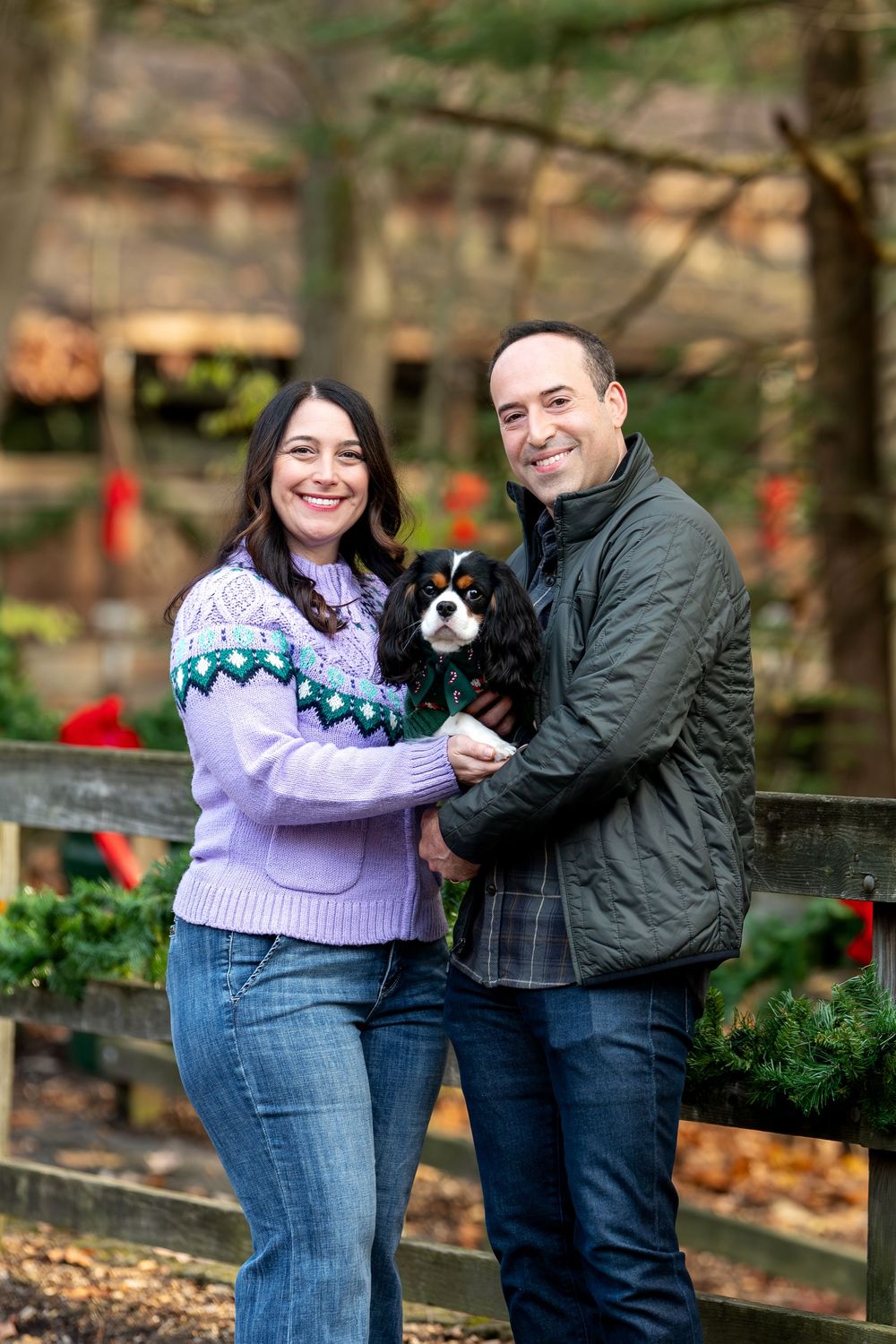 Couple posing with Cavalier King Charles Spaniel along holiday-decorated wooden fence outdoors.