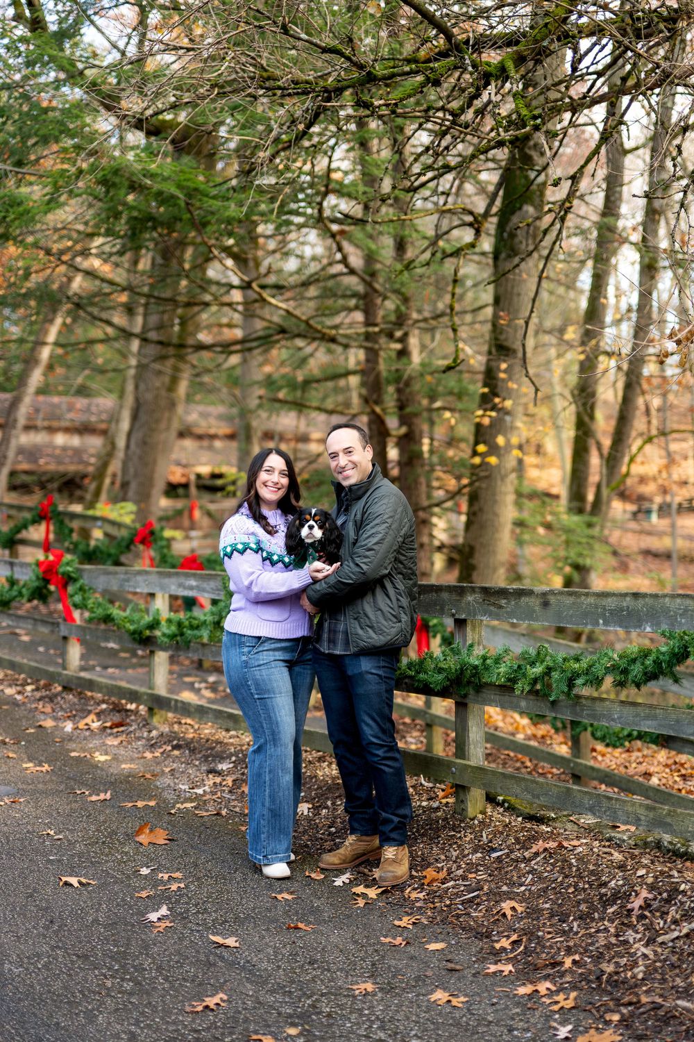 Couple holding a Cavalier King Charles Spaniel near a Christmas wreath-decorated fence in an autumn forest.