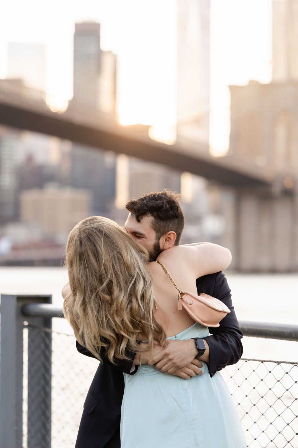 A romantic couple embraces and kisses against a blurred urban backdrop with a bridge in the distance.