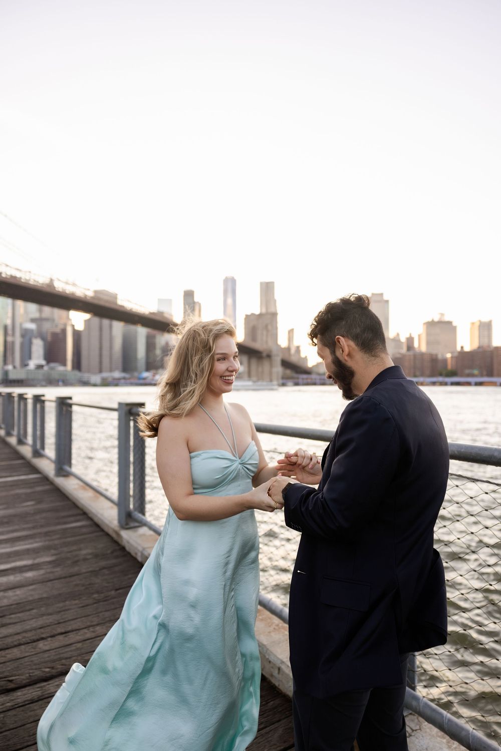 A couple in formal attire shares a romantic moment on a waterfront pier with the city skyline in the background.
