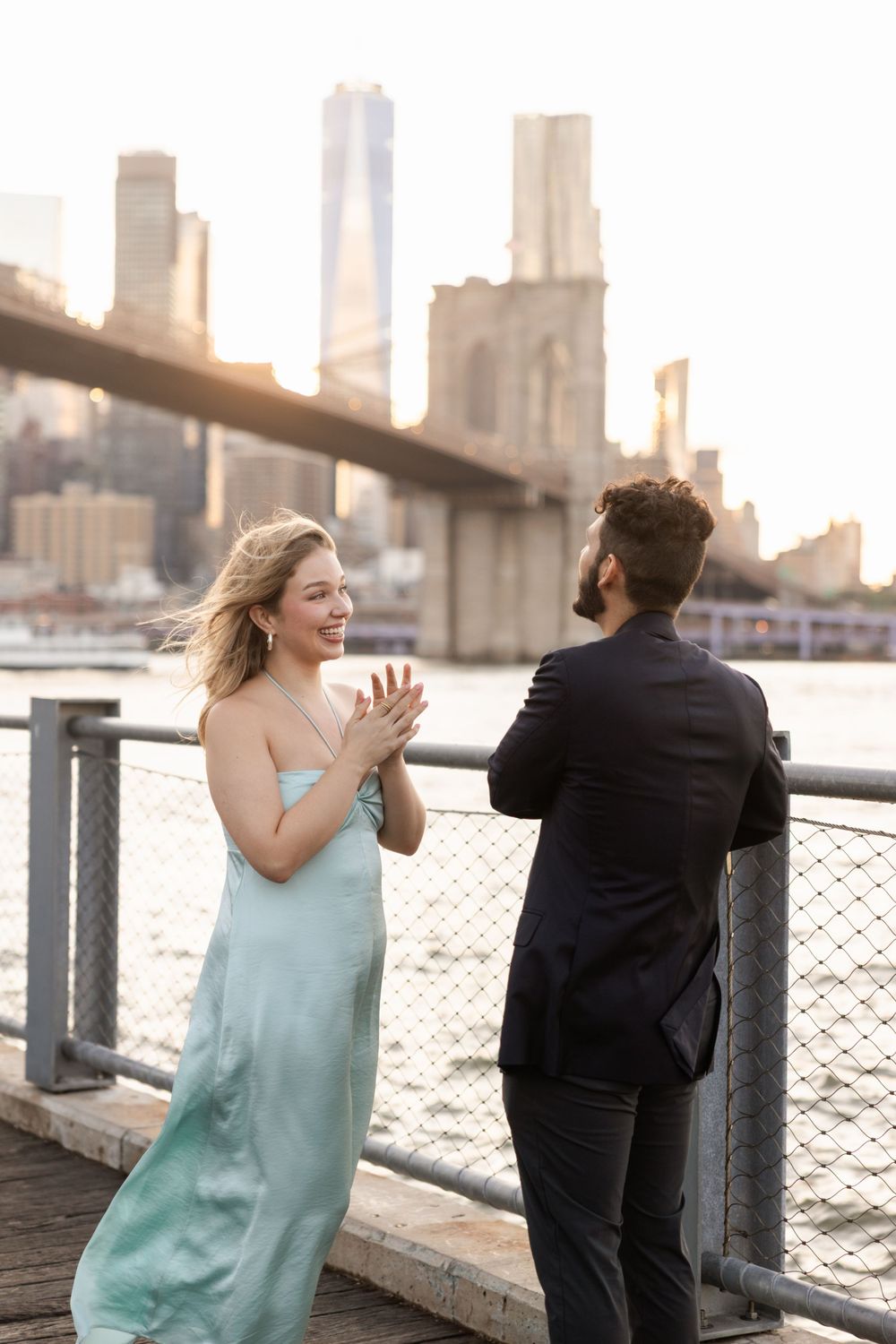 A couple in formal wear playfully interacts along a waterfront railing with the Brooklyn Bridge and city skyline at sunset.