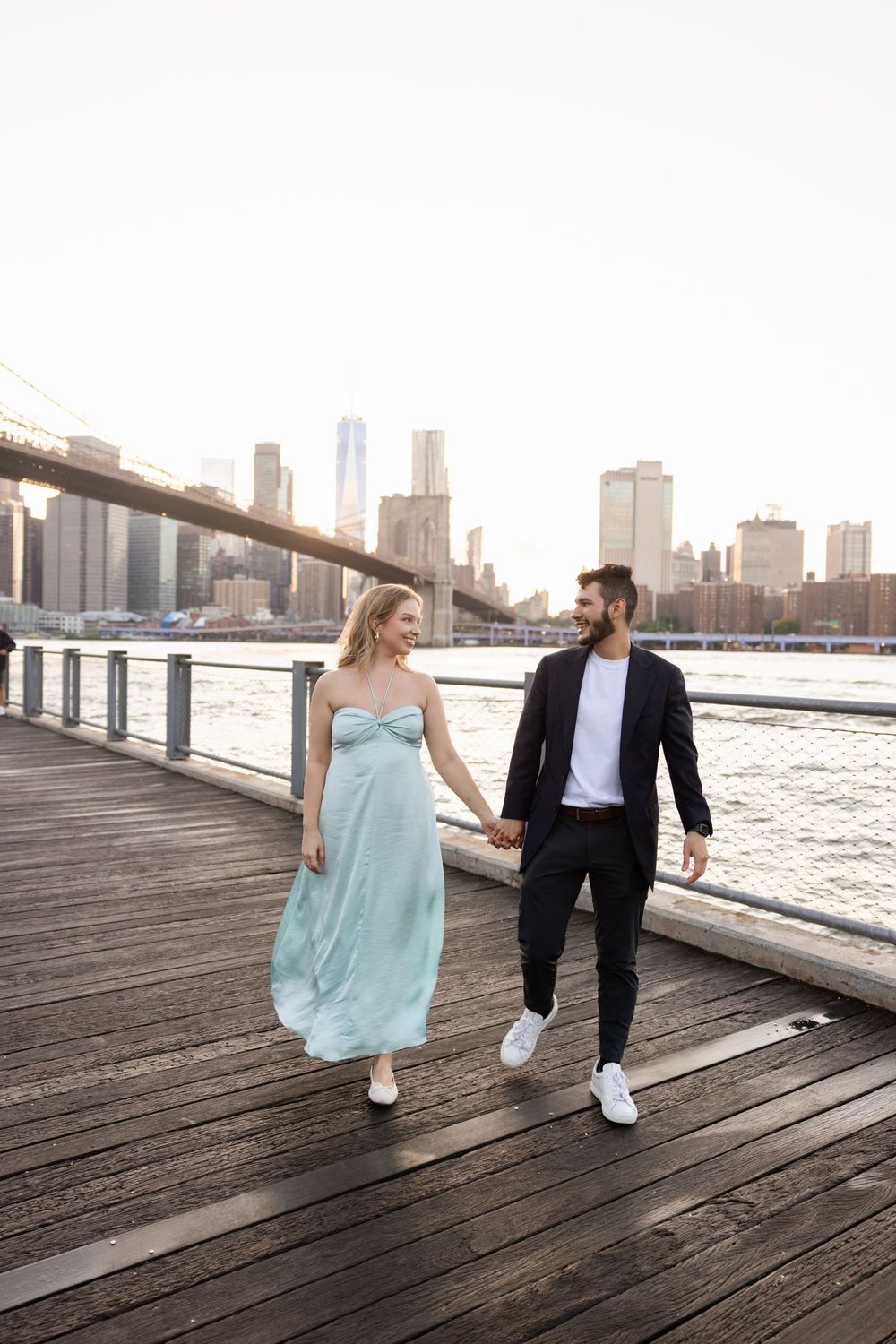 Couple walking along a waterfront boardwalk with the Brooklyn Bridge and Manhattan skyline in the background.
