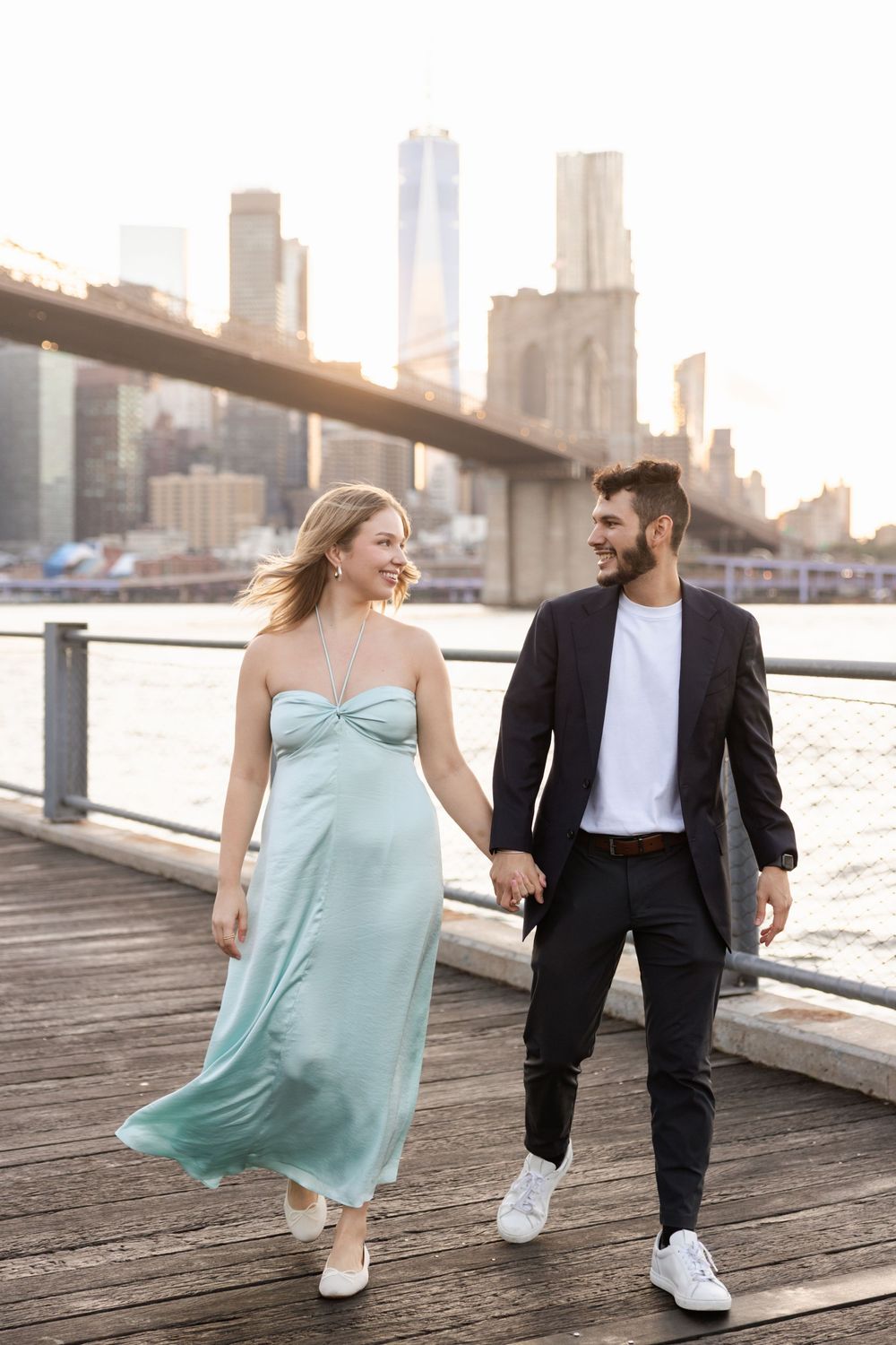 Couple in contrasting outfits walk along wooden pier with NYC skyline in background at sunset.