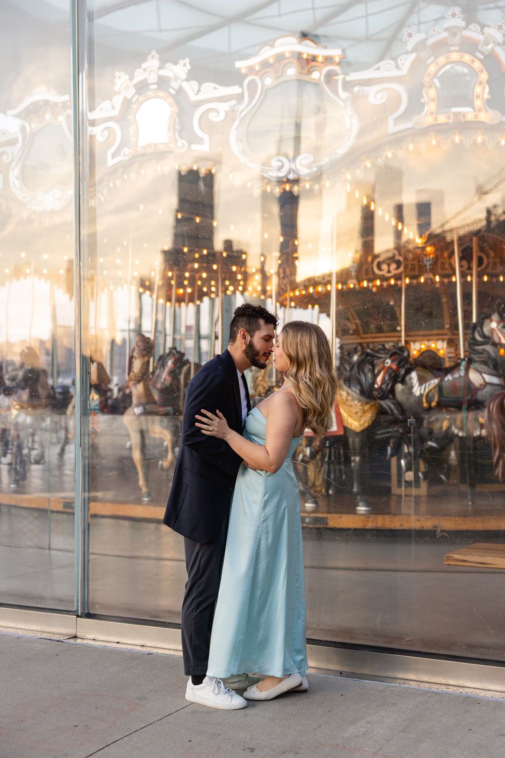 A couple embraces in front of an illuminated vintage carousel at dusk, with twinkling lights creating a romantic atmosphere.