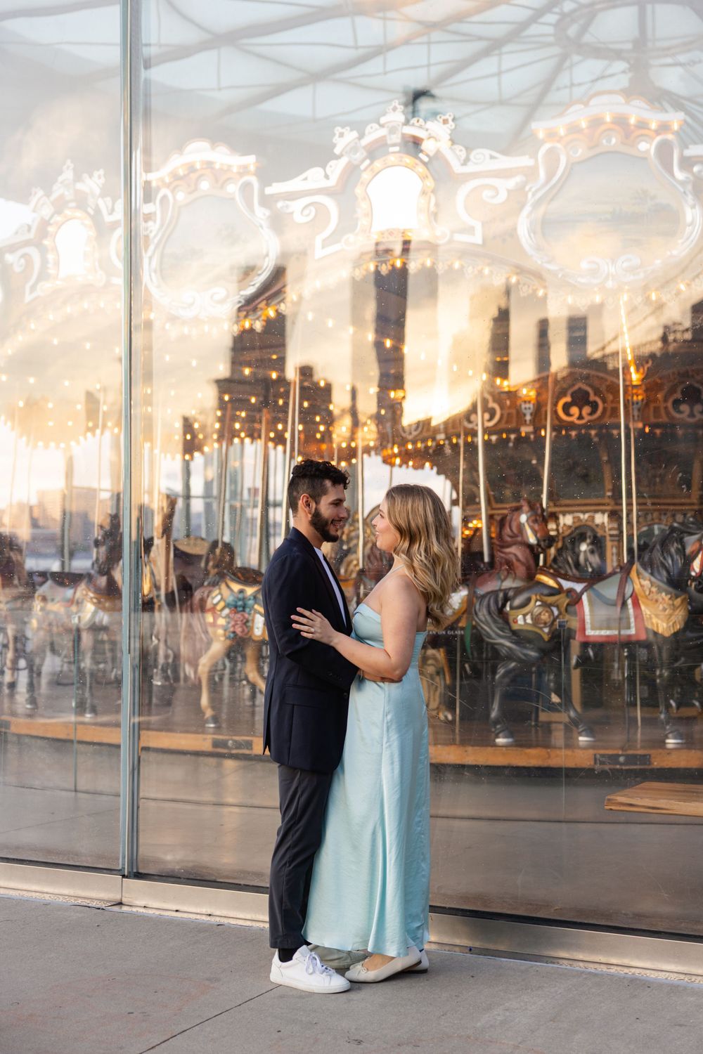A romantic couple shares an intimate moment in front of an illuminated carousel at sunset.