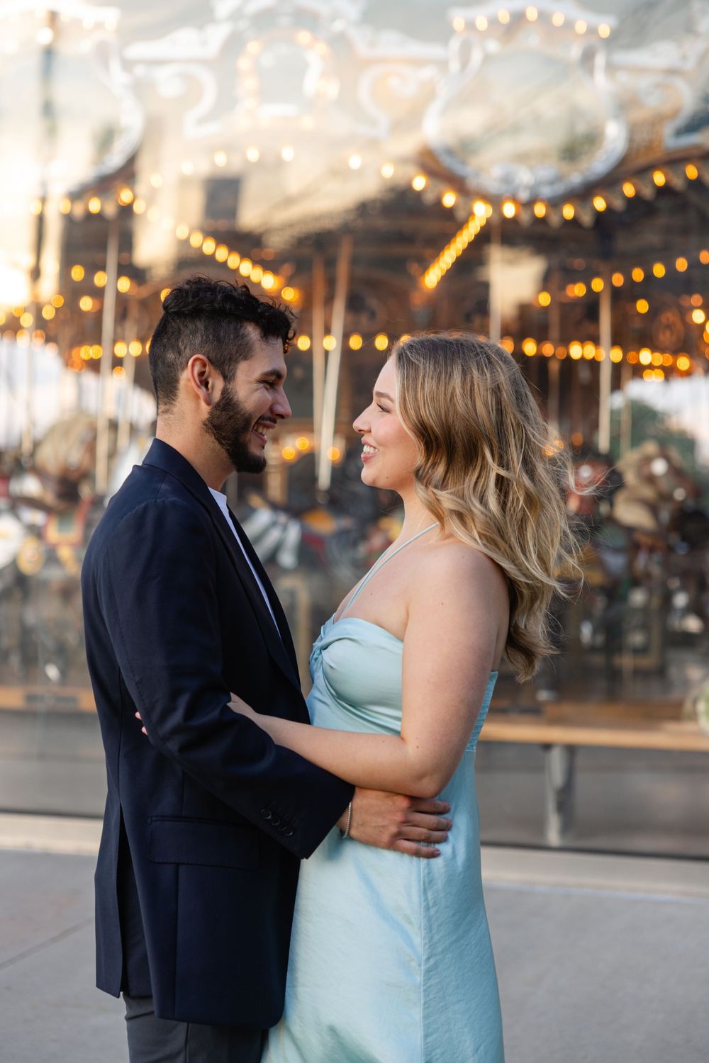 A couple shares a romantic moment in front of a vintage carousel with twinkling lights at dusk.