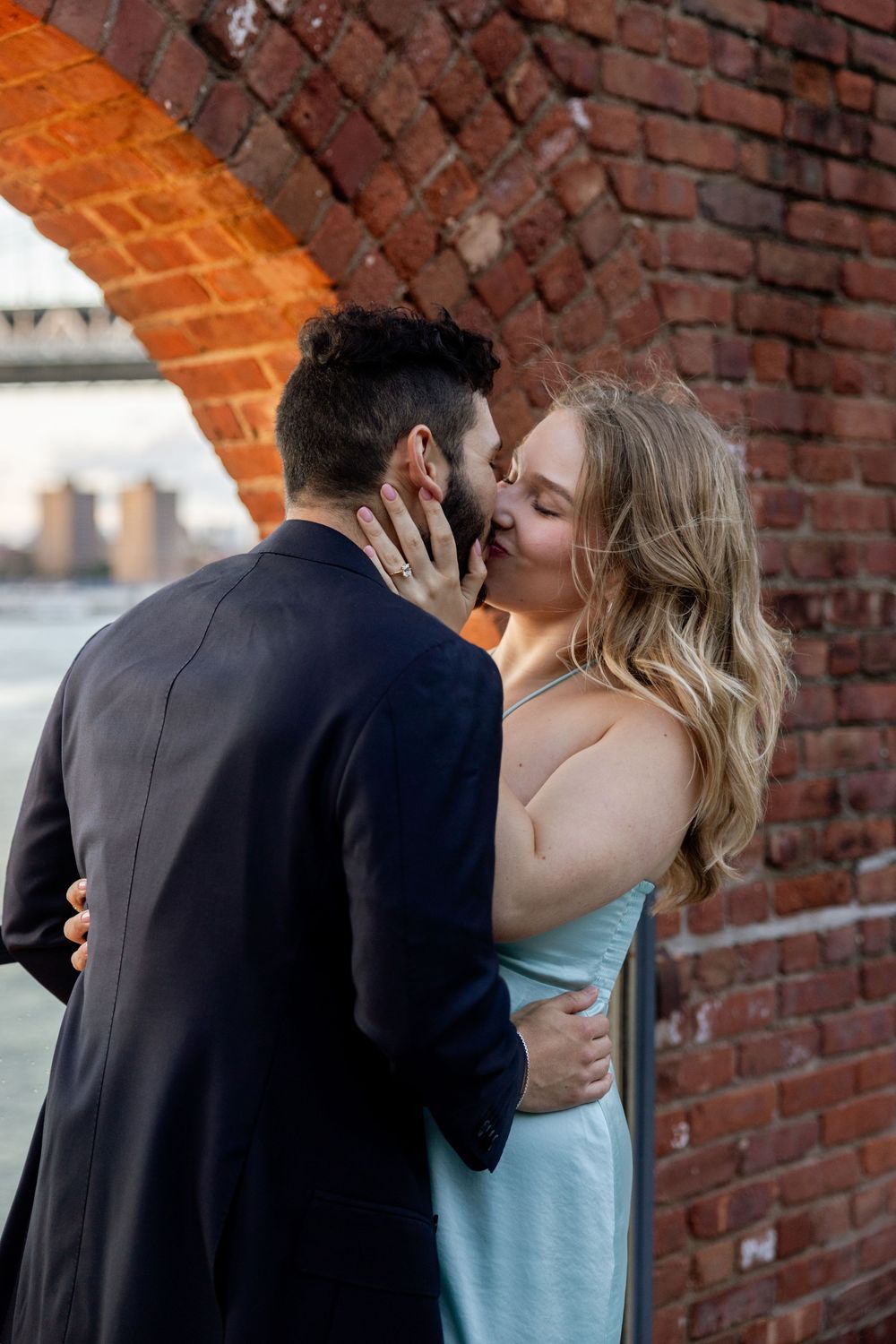 A couple embraces and kisses against a rustic brick wall backdrop with arched details.