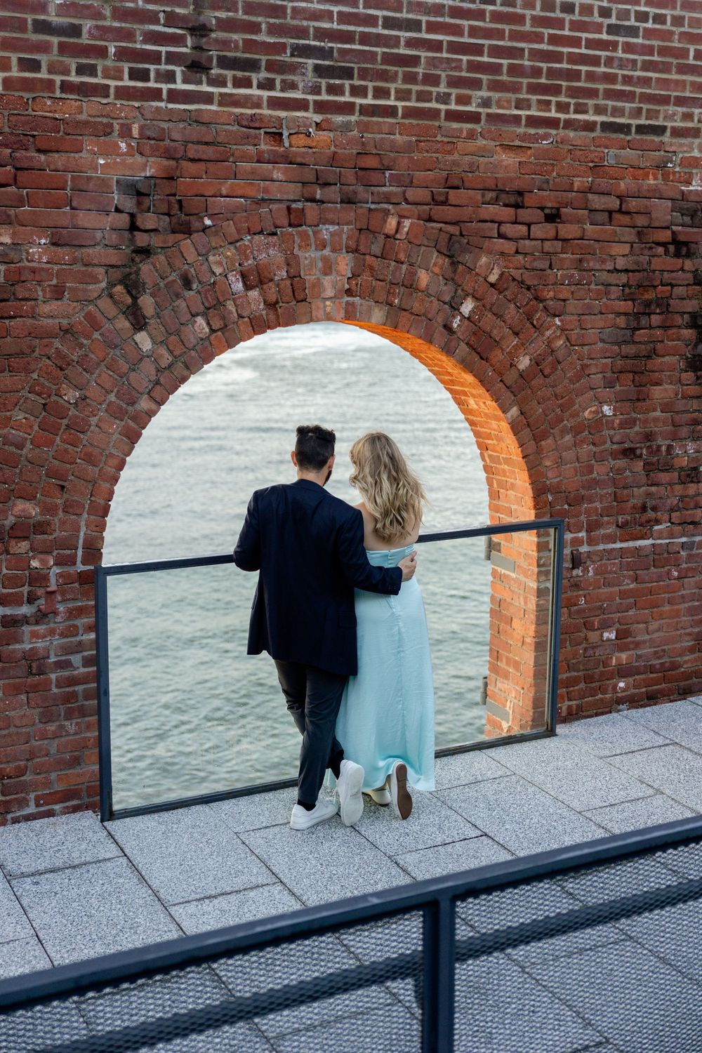 A couple walks through a brick archway along a stone path with a metal railing in the foreground.