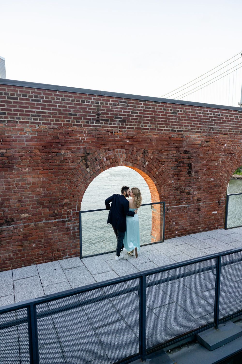 A couple playfully peeks through arched openings in a long brick wall with metal railings.