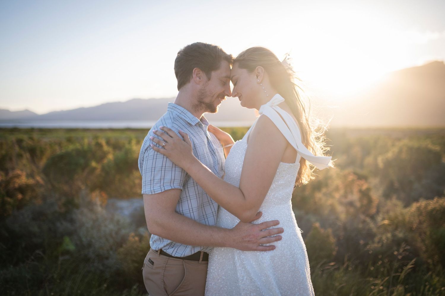 Romantic couple embraces in golden sunlight against scenic mountain landscape during sunset photo session.