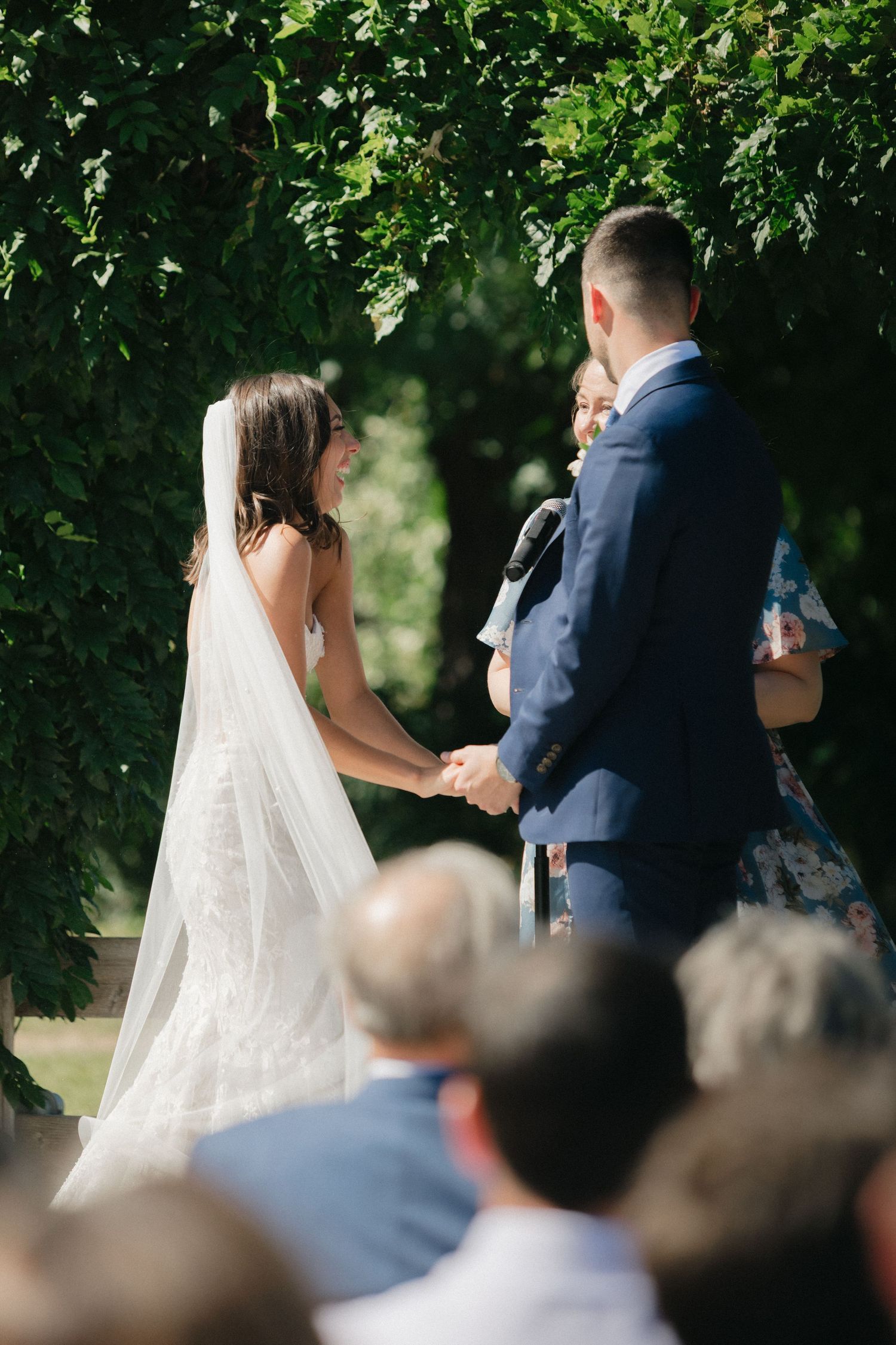 A couple exchanges vows during an outdoor wedding ceremony surrounded by lush green trees and seated guests.