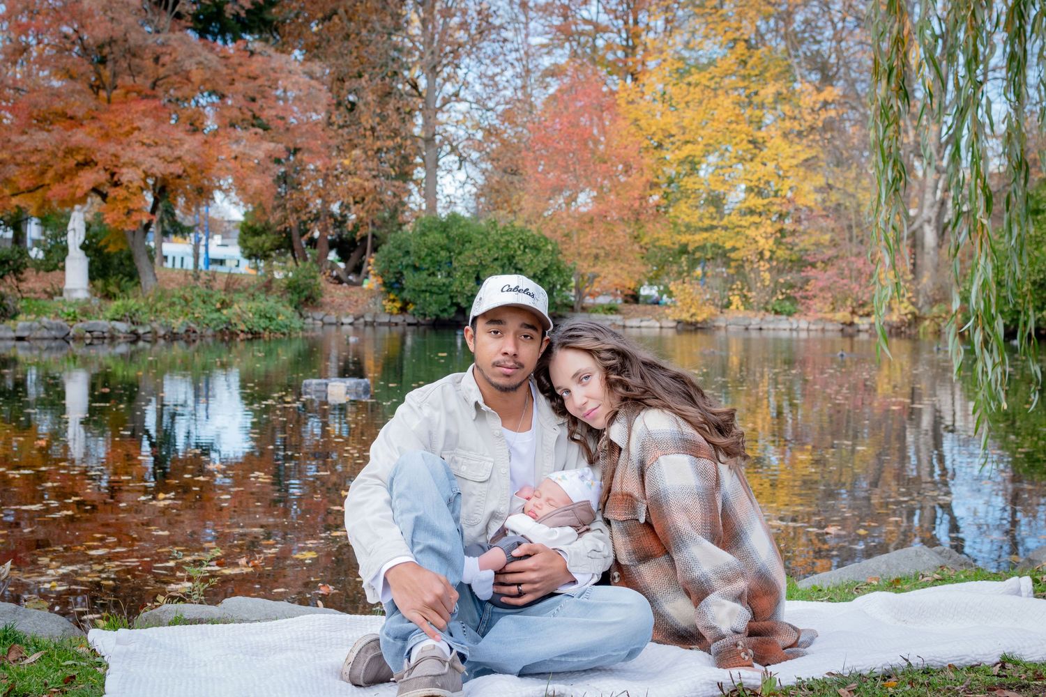 Family with a newborn sitting by the pond with autumn trees during a fall photo session at Wright Park in Tacoma.