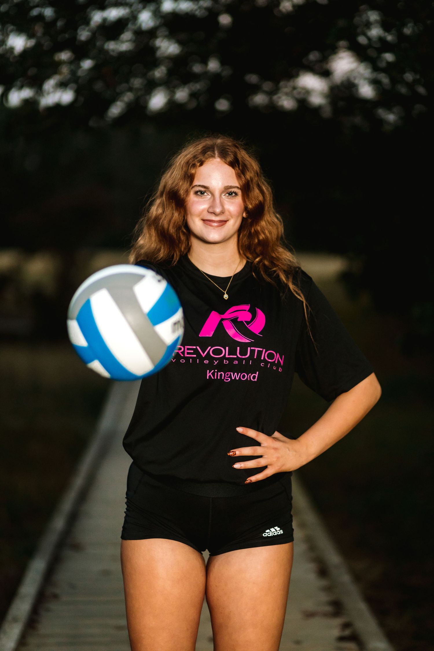 A volleyball player in a black uniform poses with a blue and white volleyball in an outdoor setting.