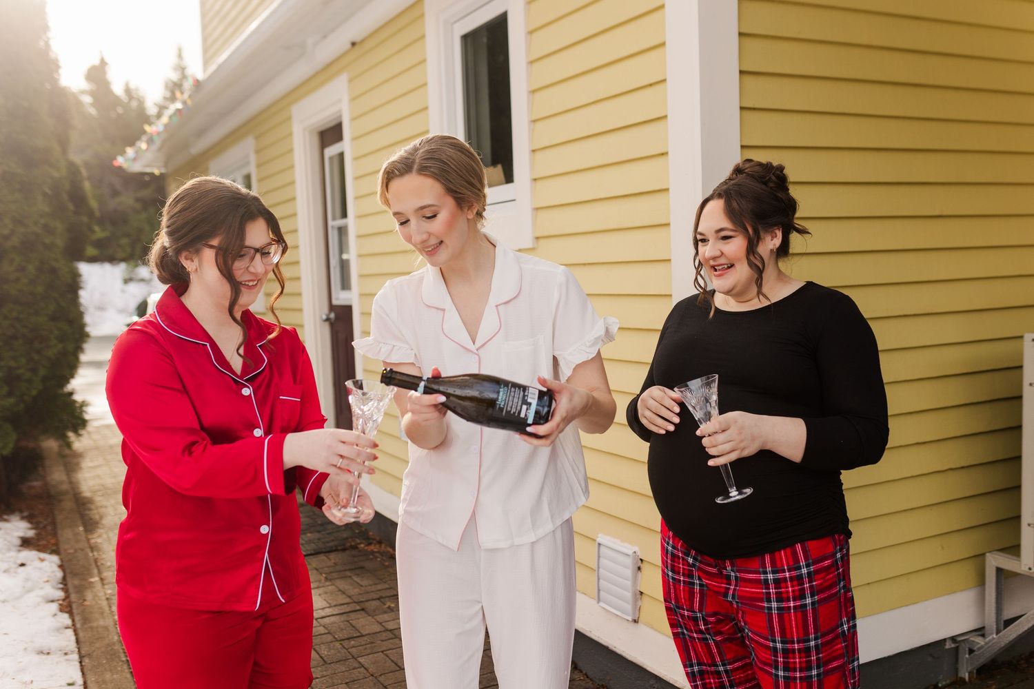 Three friends in festive winter attire share wine and laughter on a porch beside a yellow house.
