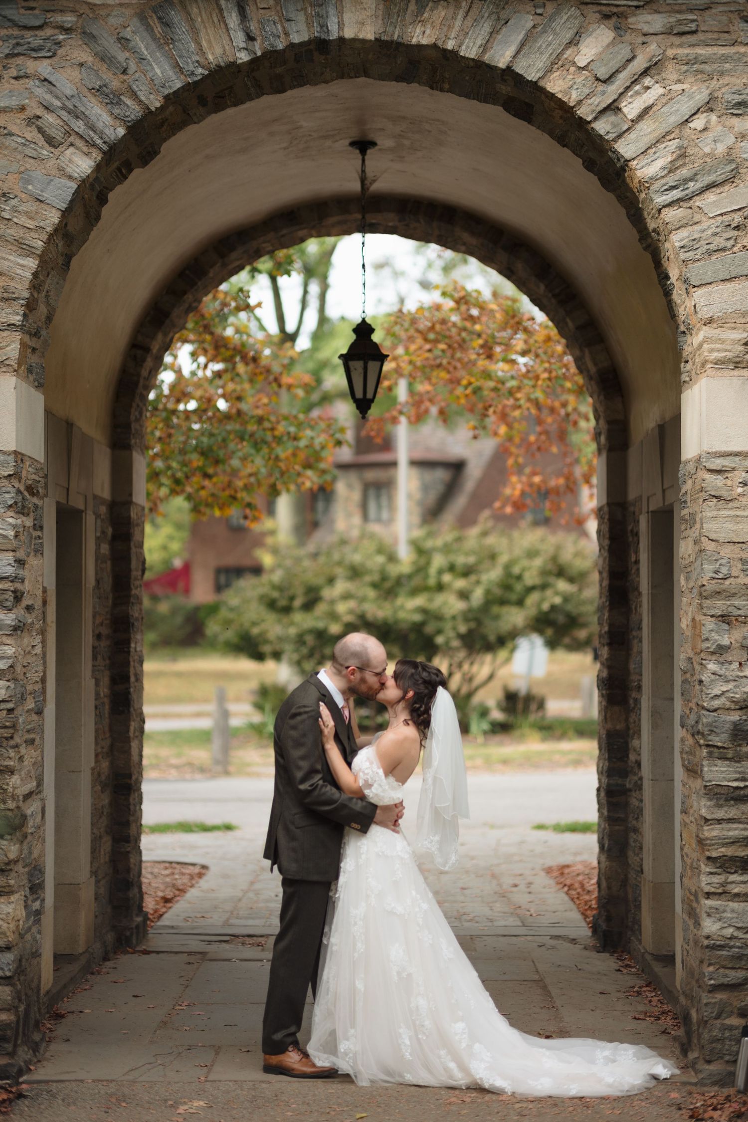 A romantic wedding couple shares intimate moments under a stone archway surrounded by autumn foliage.