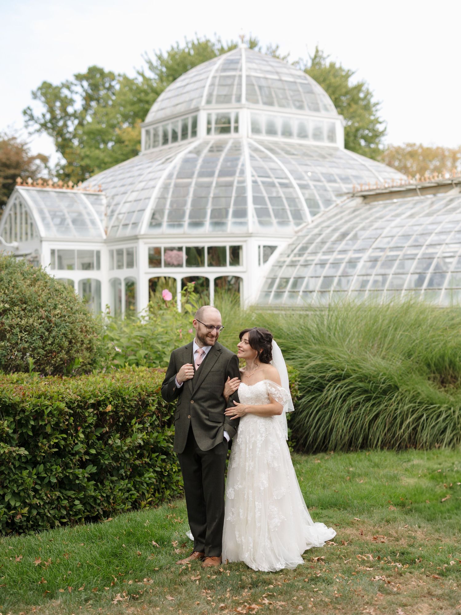 A couple poses together in front of elegant glass greenhouse conservatory structures in a garden setting.