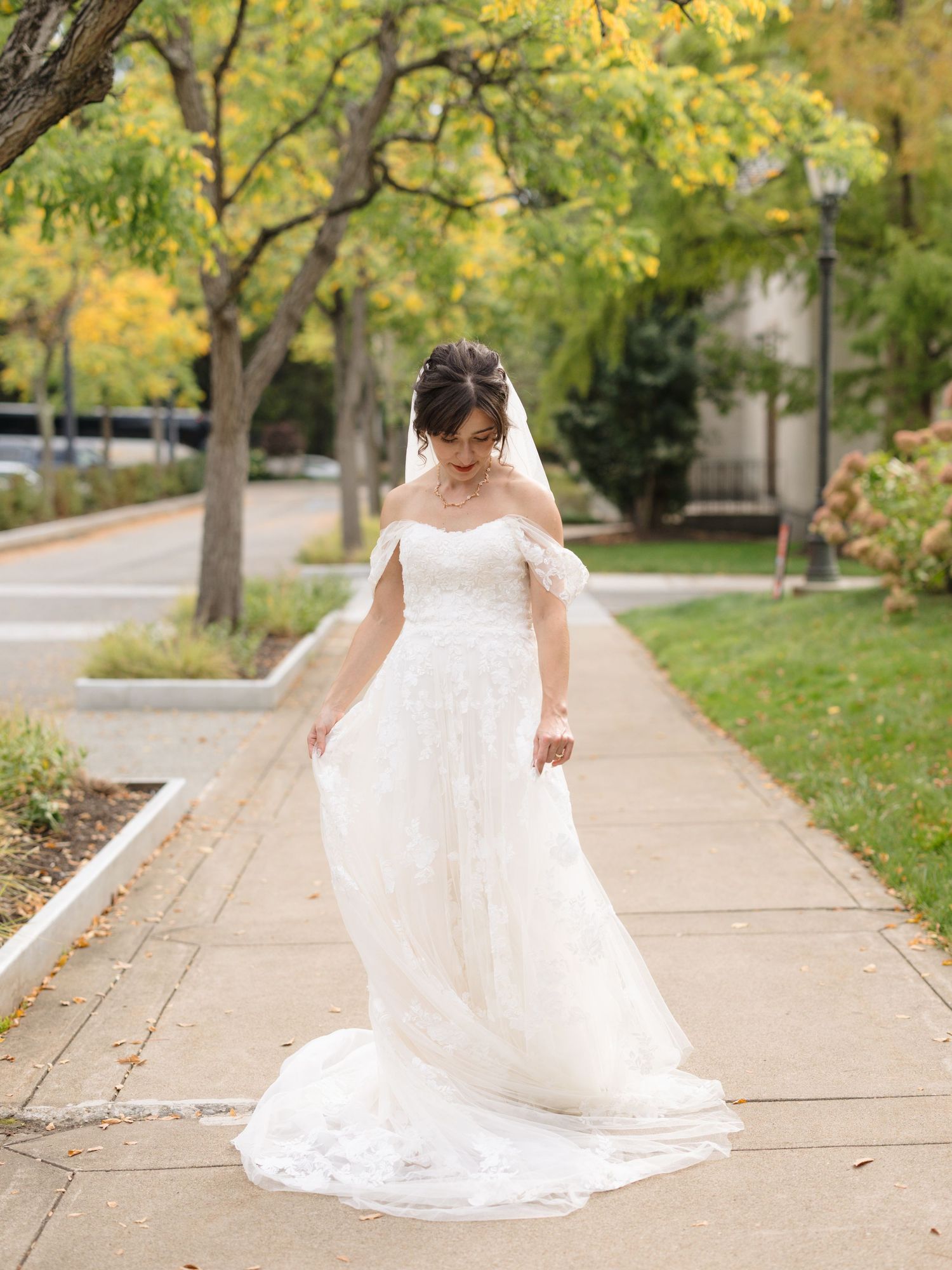 A bride in an elegant off-shoulder white gown twirls on a tree-lined sidewalk during autumn.