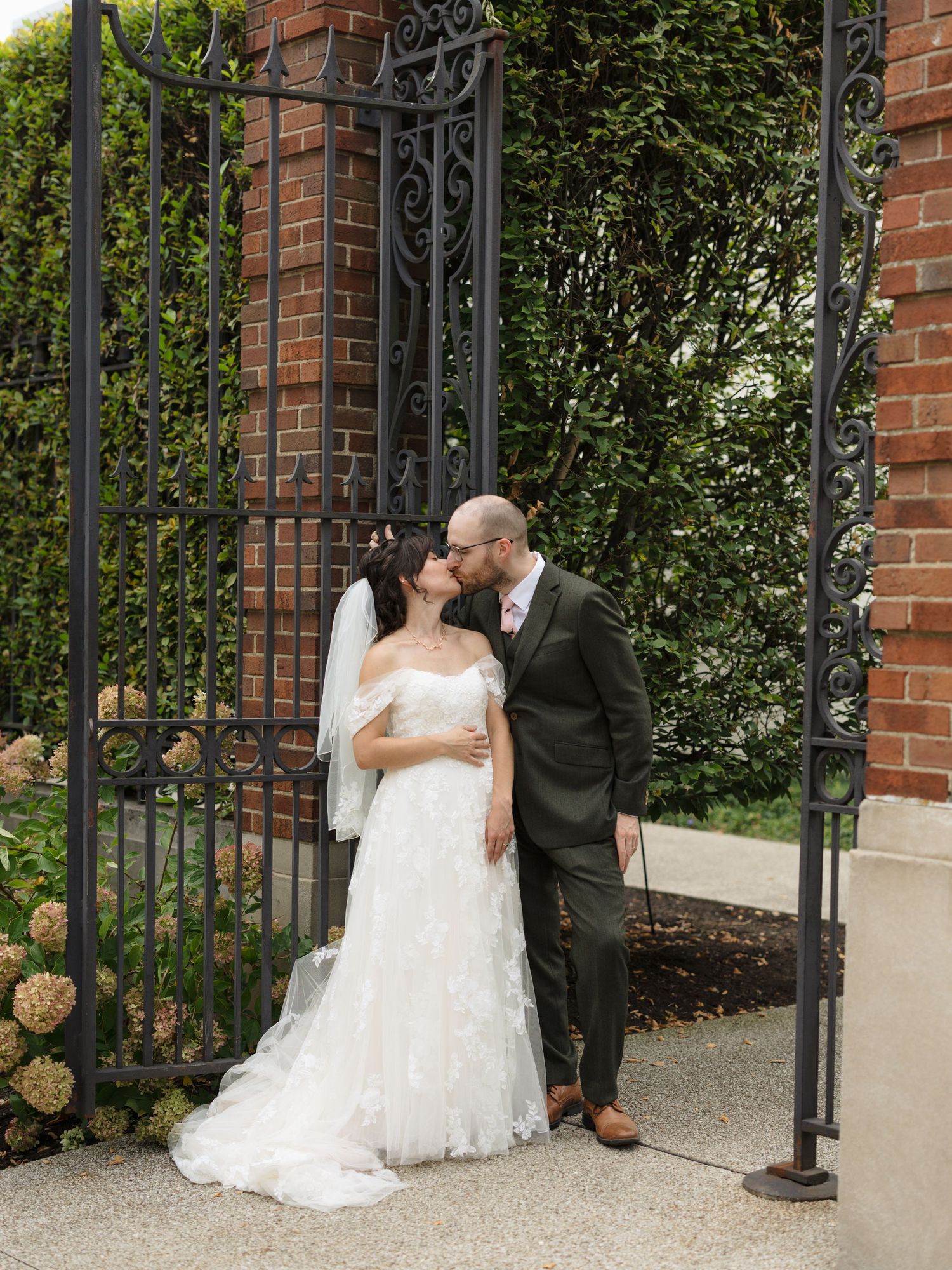 A couple shares an intimate moment by ornate iron gates and brick pillars in a garden setting.