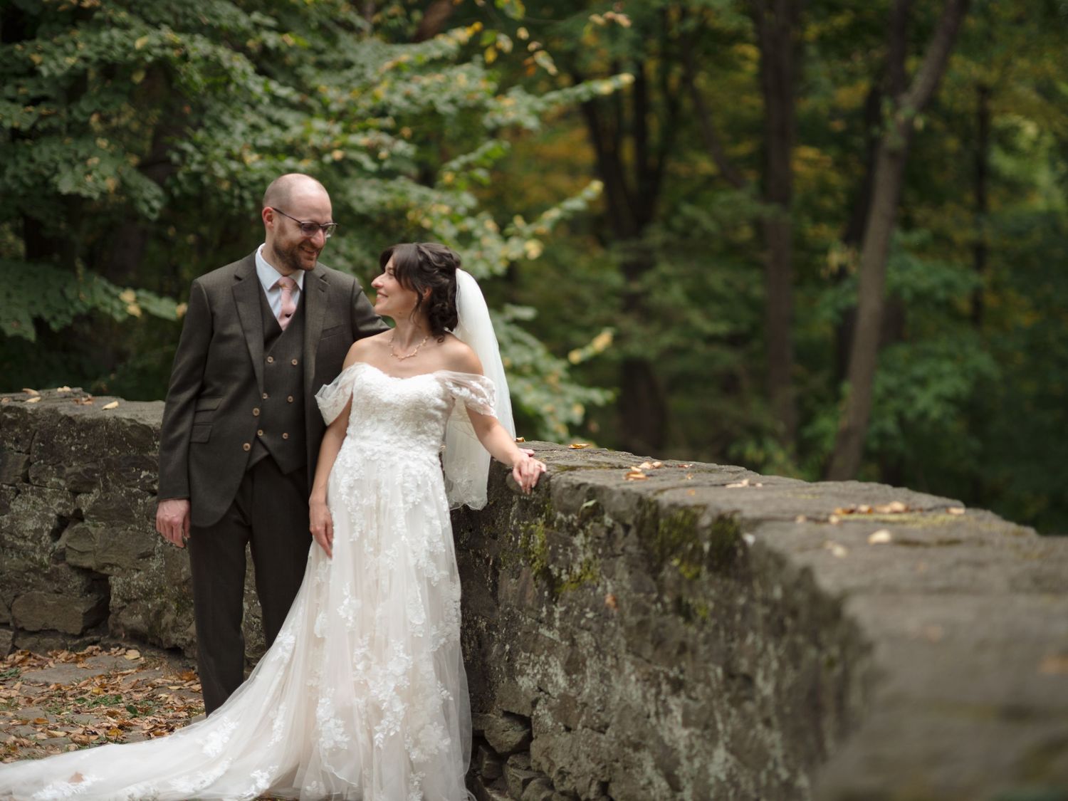 A couple in wedding attire pose together on stone steps surrounded by lush green foliage in a garden setting.