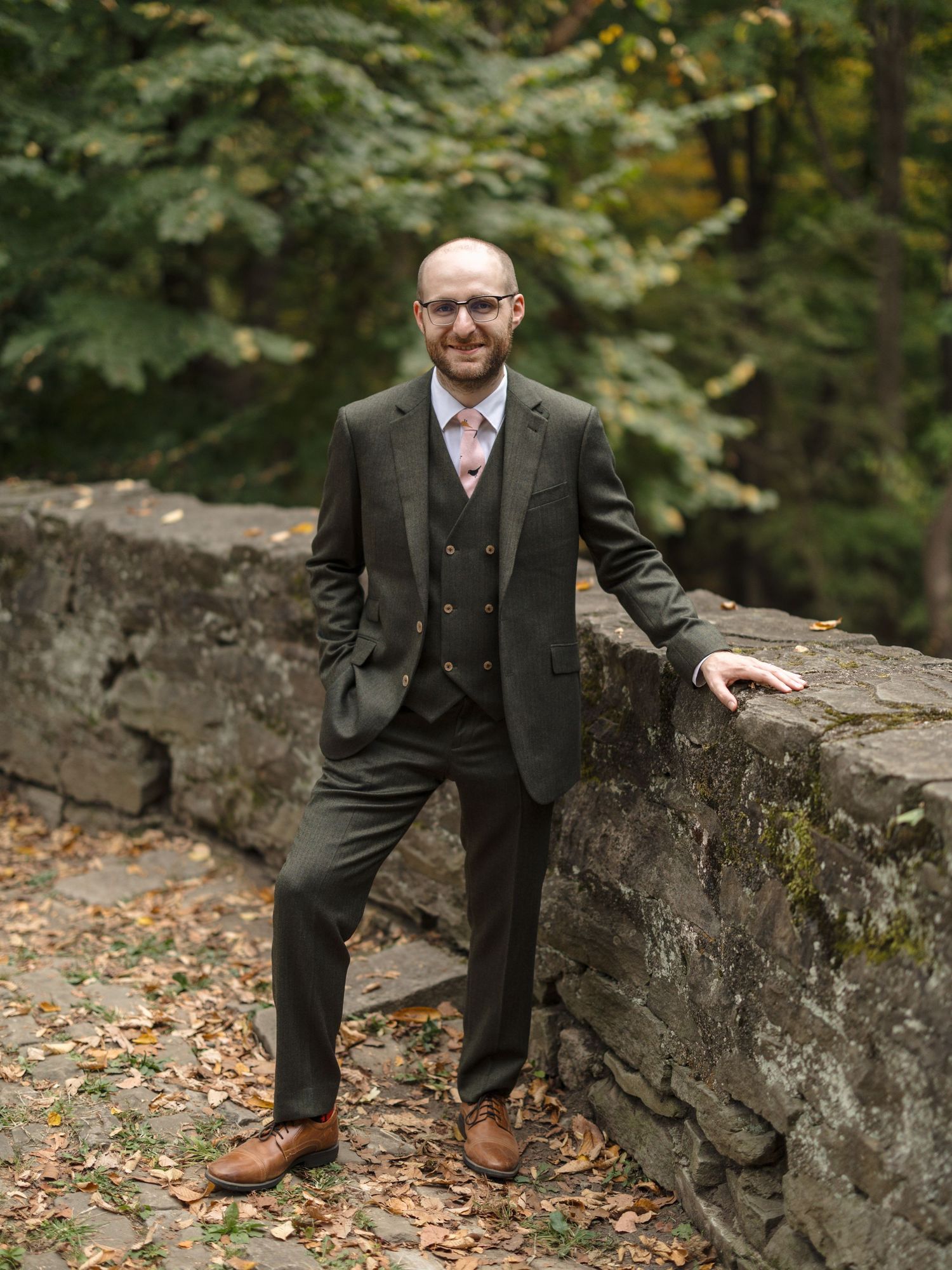Professional portrait session featuring a three-piece olive suit and brown shoes against a rustic stone wall backdrop.