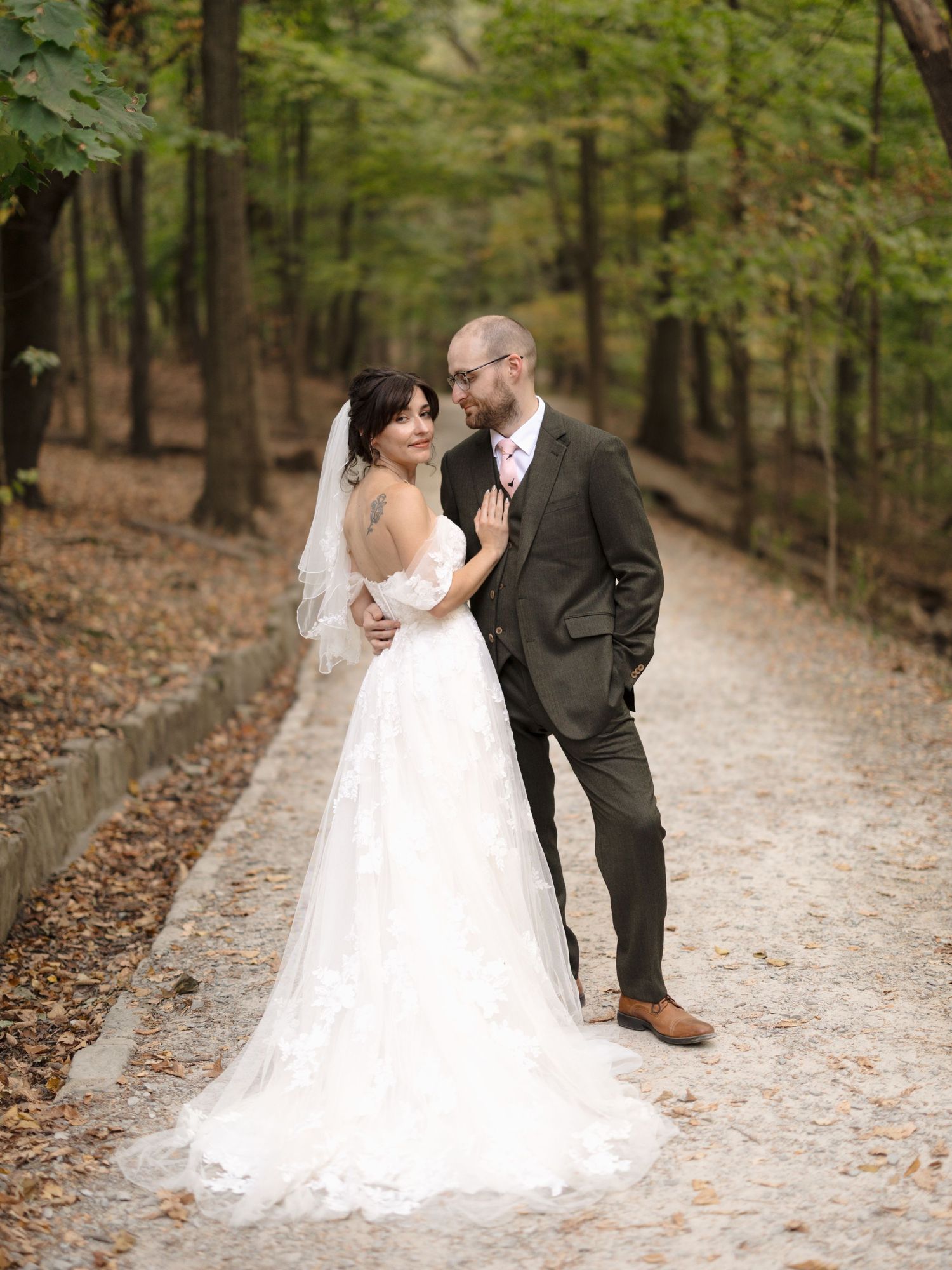 A romantic wedding couple poses on a leaf-covered forest path in autumn, with the bride's long veil trailing behind.