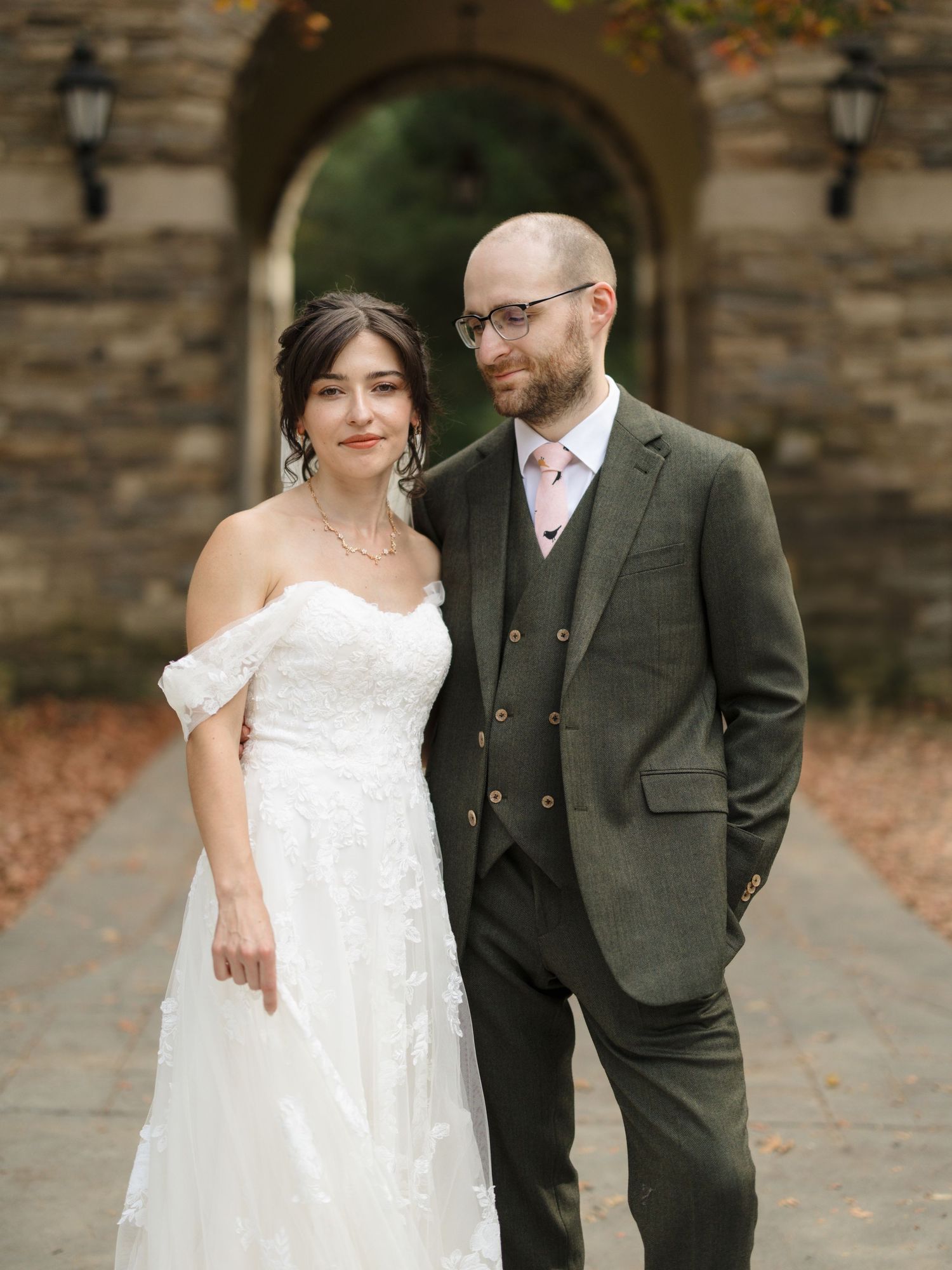 A couple in wedding attire walking together outdoors, bride in white lace dress and groom in olive green suit.