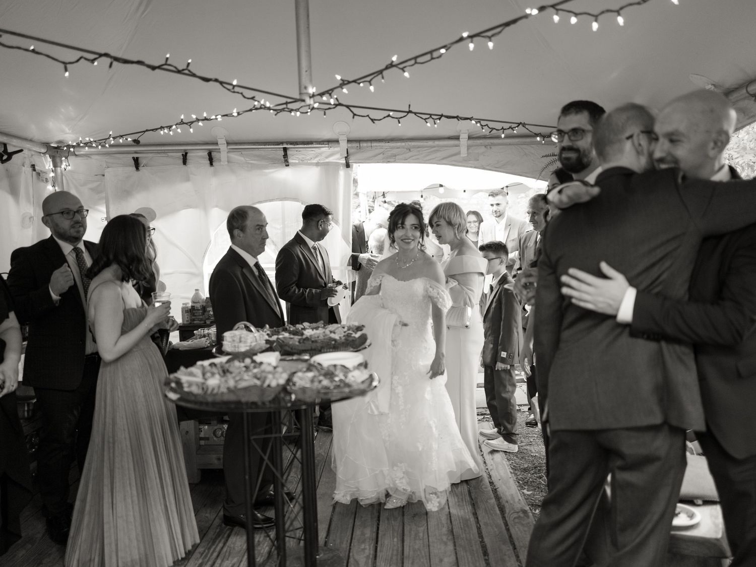 Black and white photo of wedding reception guests mingling under string lights in a tented outdoor venue.