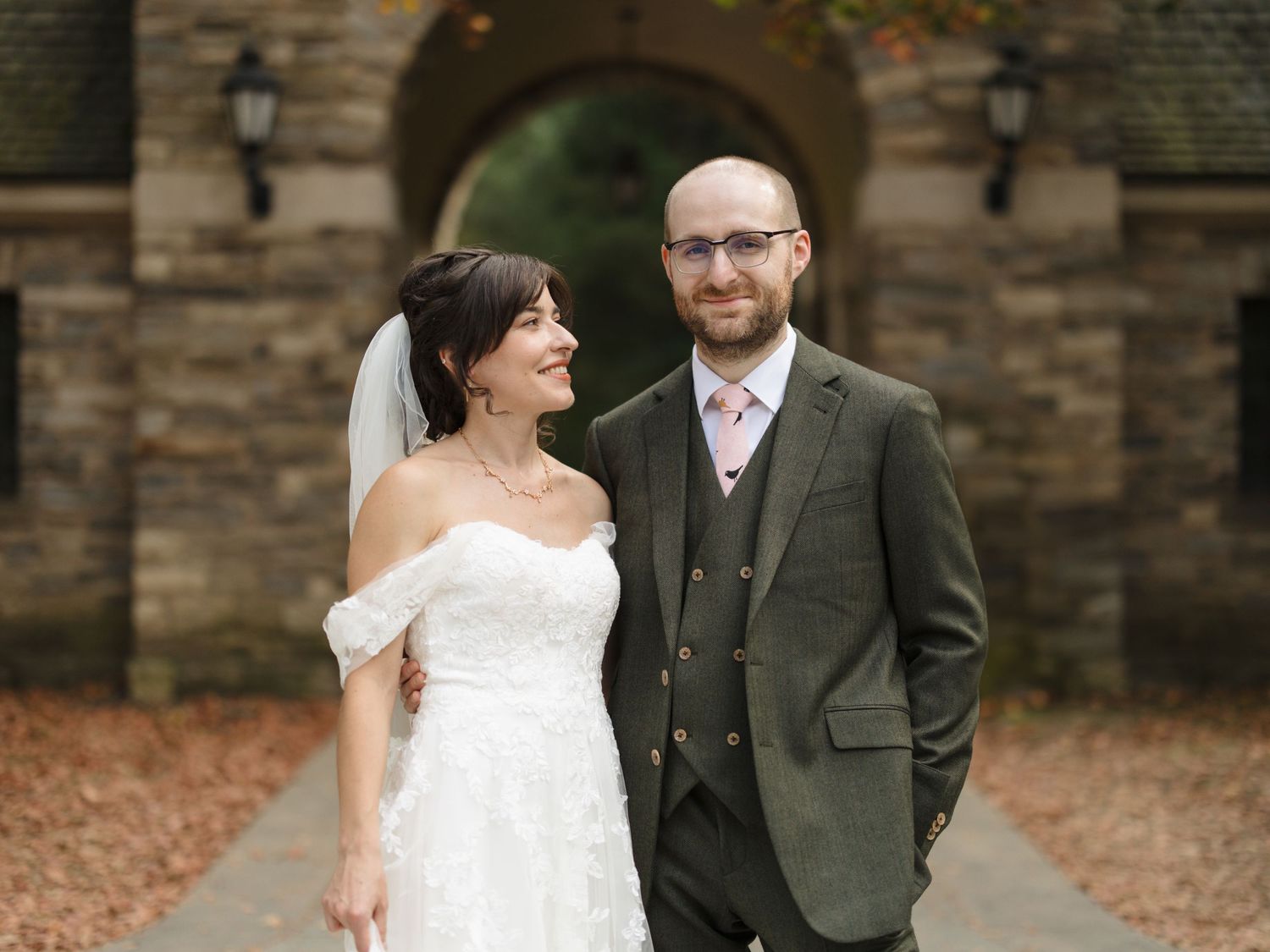 A bride in an off-shoulder white dress and groom in a gray suit stand together outside a stone building on their wedding day.