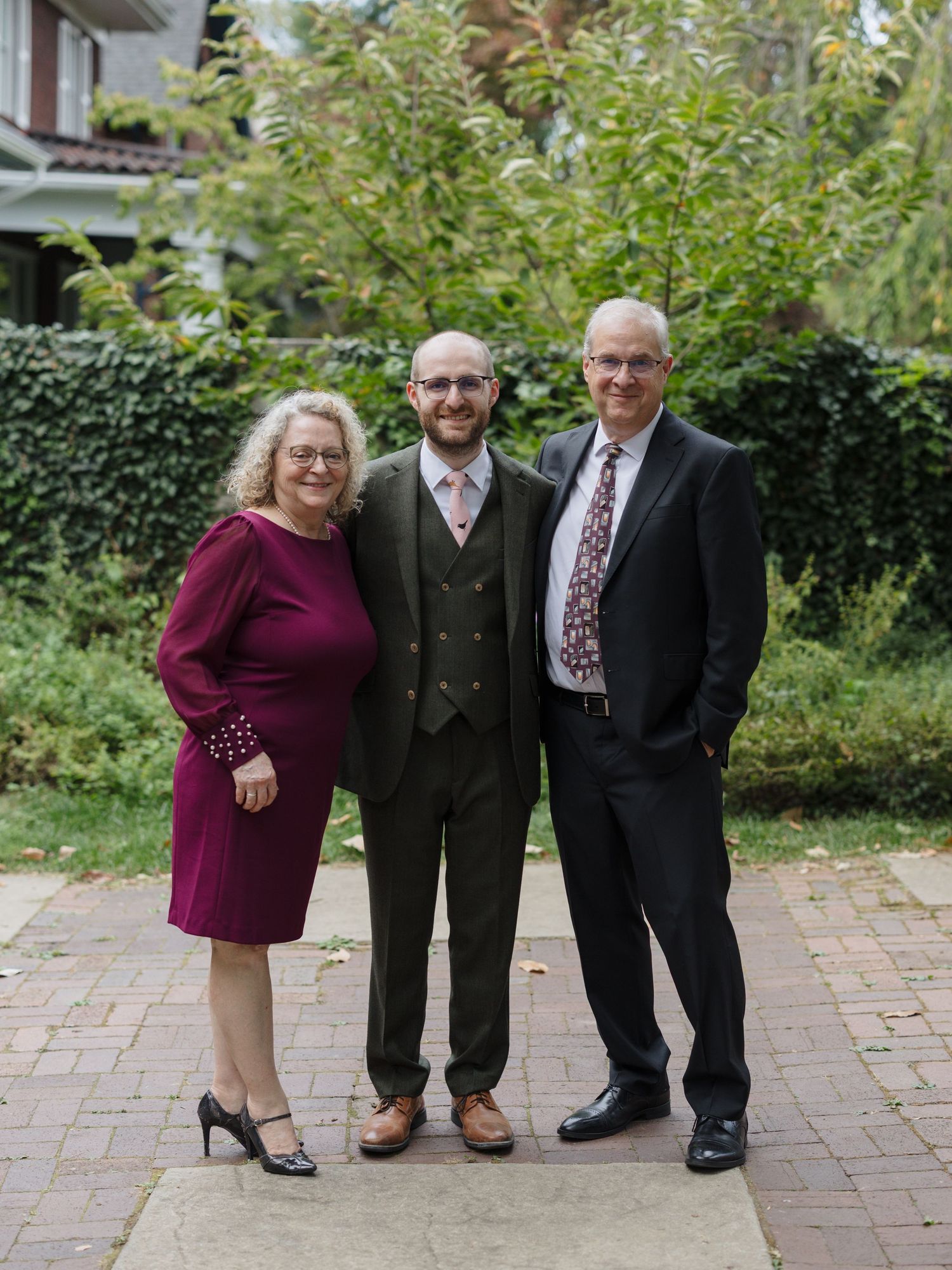 Wedding guests in formal attire pose together on a brick patio with garden greenery in the background.