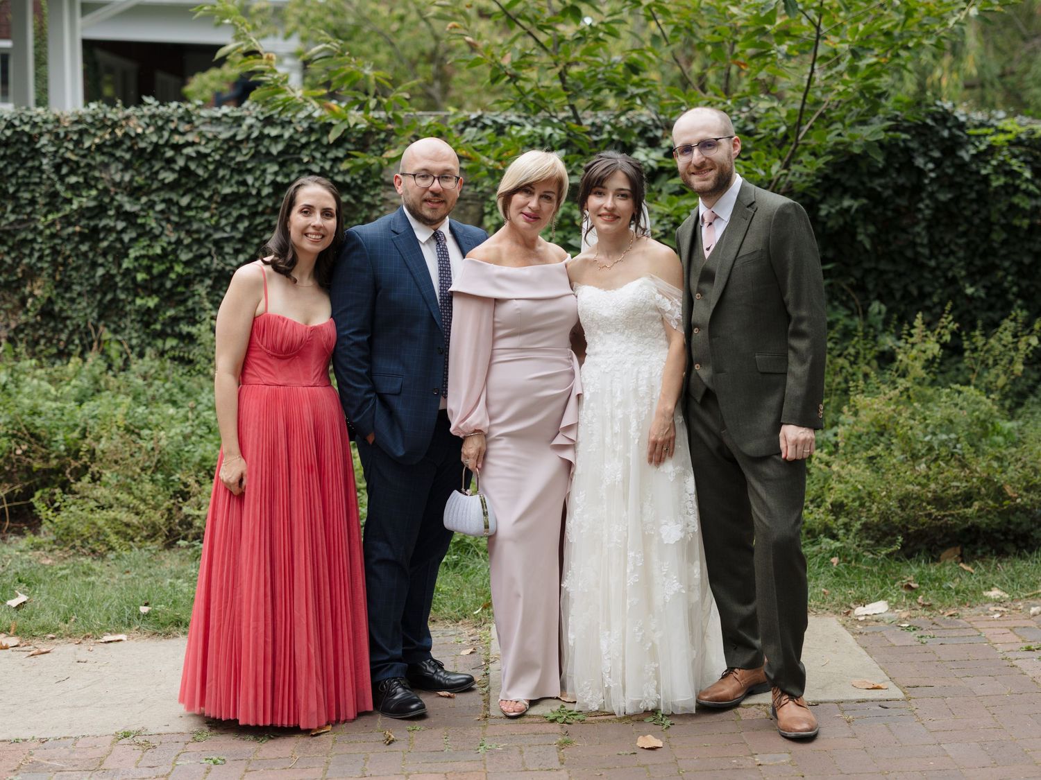 Wedding party members in coral and neutral dresses pose together for formal photos in a garden setting.