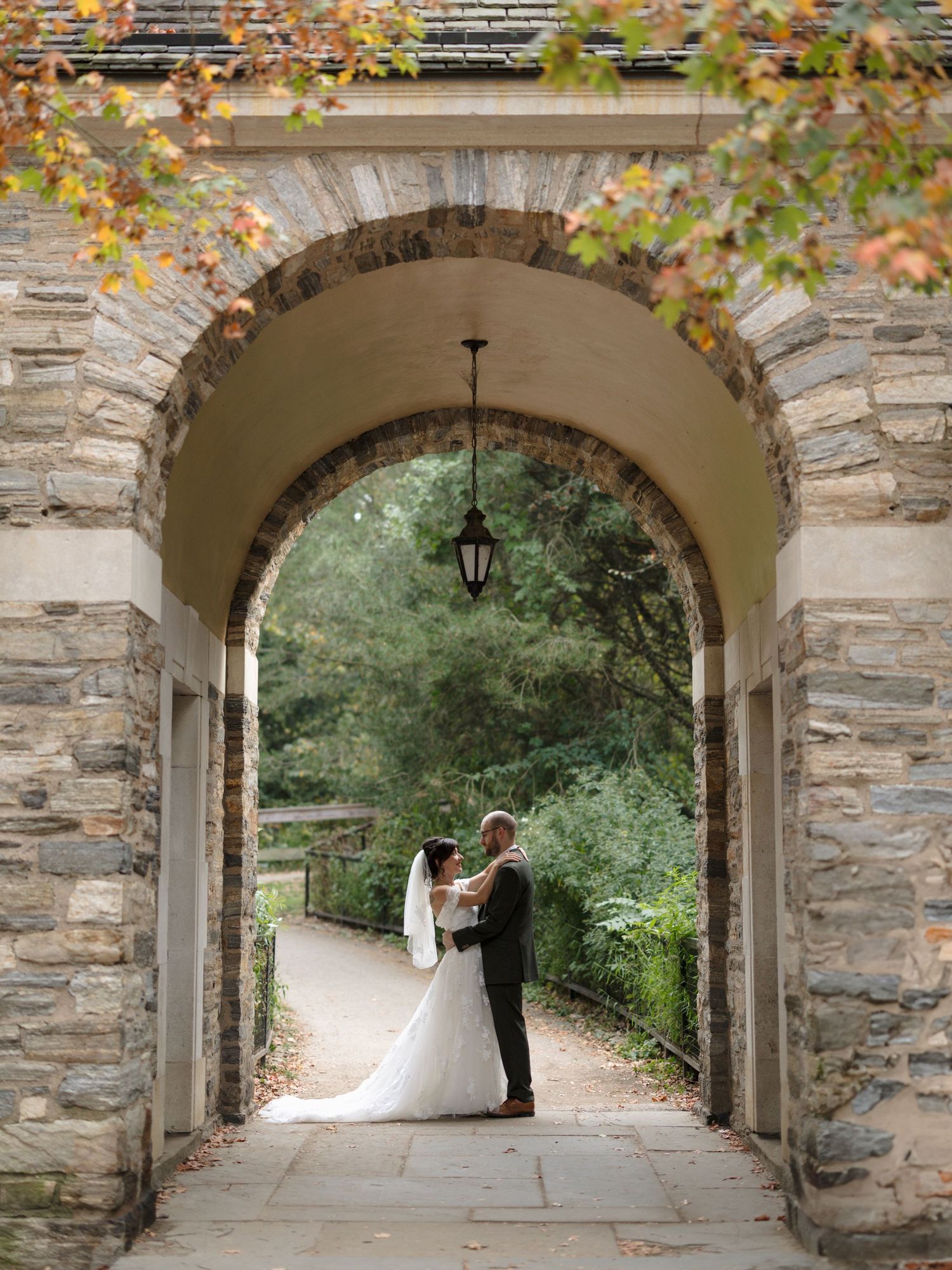 A romantic wedding couple stands under a stone archway with autumn foliage and a hanging lantern.