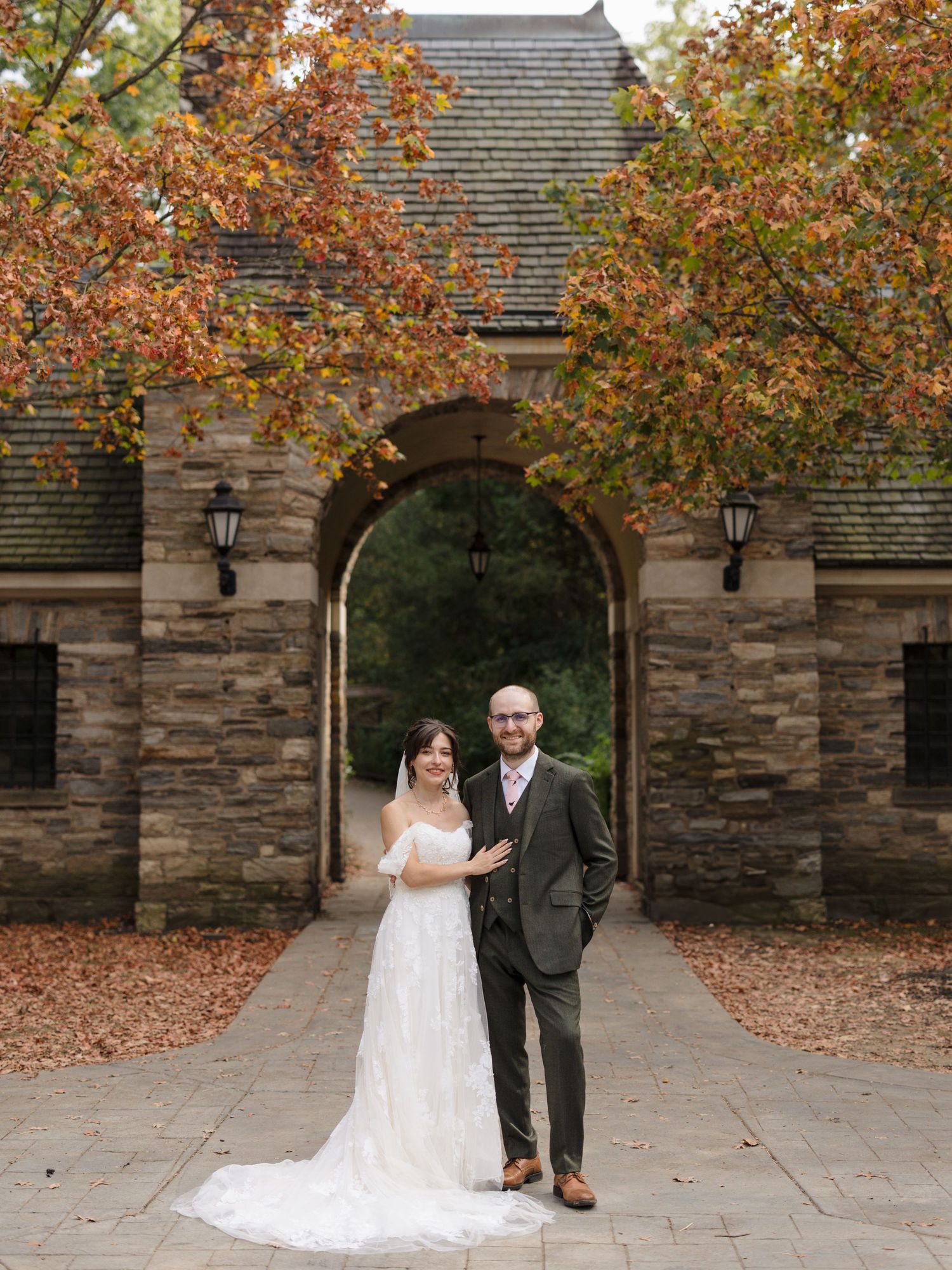 A wedding couple stands in front of a stone archway surrounded by autumn foliage at an elegant venue.