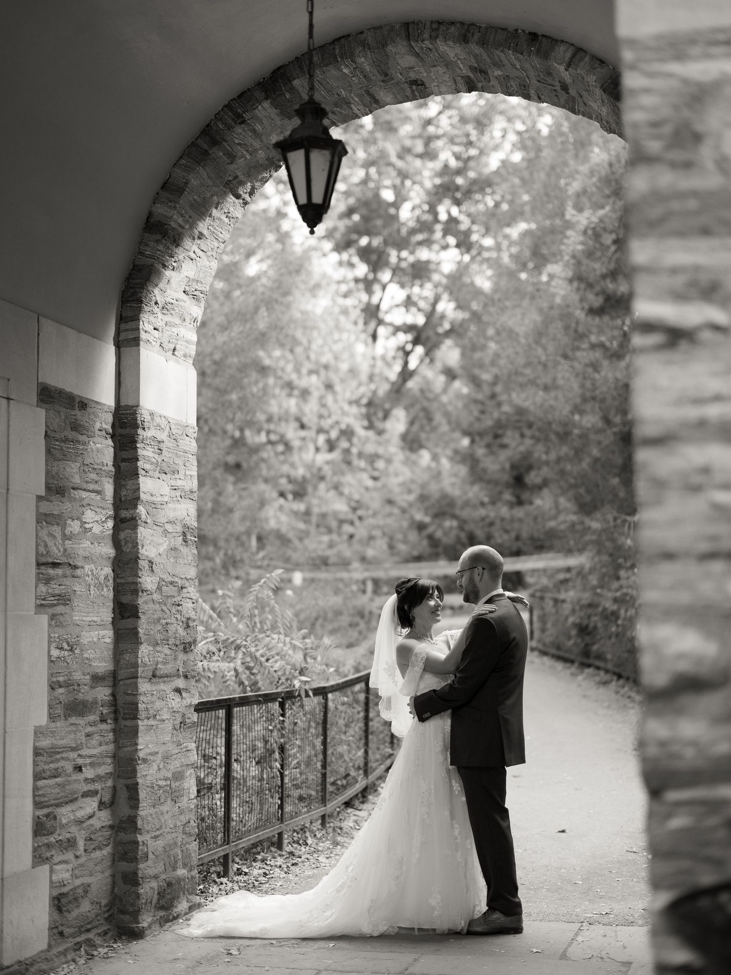 A romantic black and white wedding photo of a couple embracing under a stone archway with a hanging lantern.