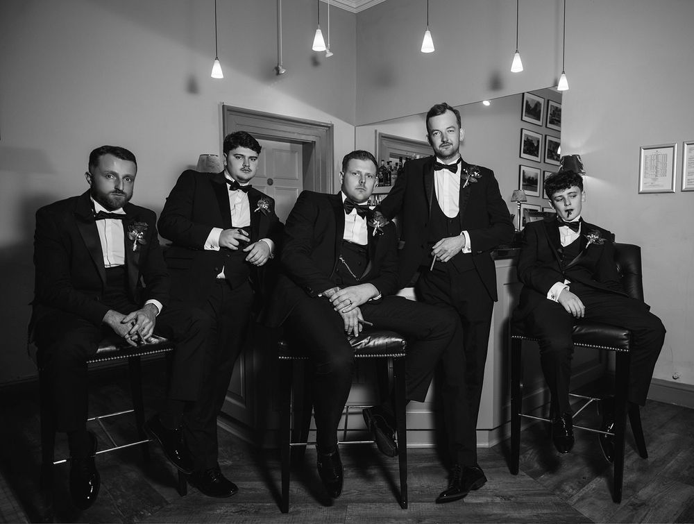 Black and white panoramic photograph showing several men in tuxedos seated in chairs along a wall with pendant lights.