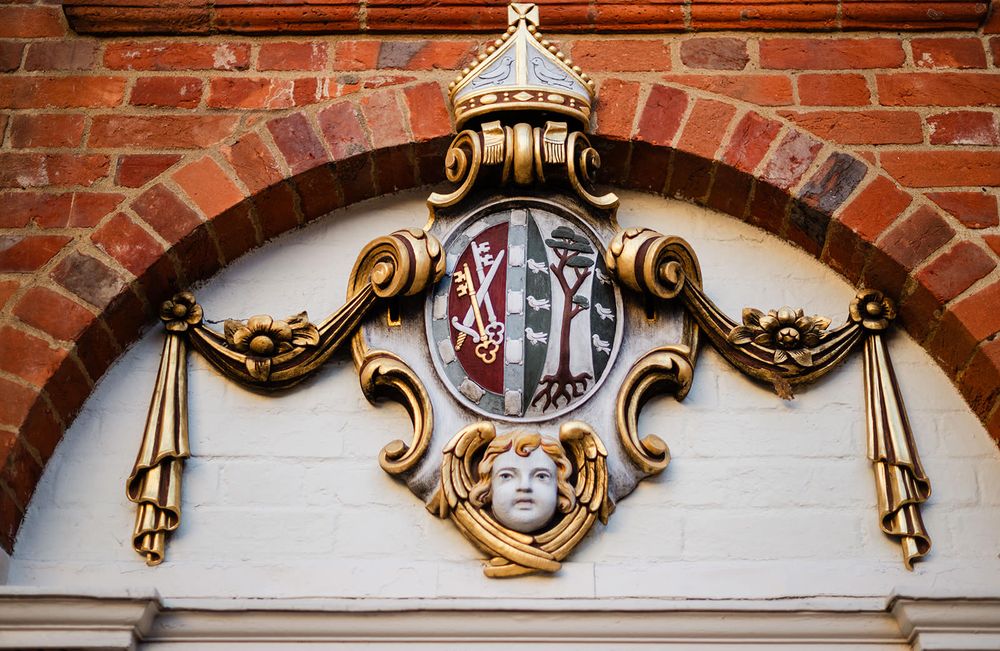 Ornate gold architectural detail with heraldic emblem mounted on red brick wall above doorway.