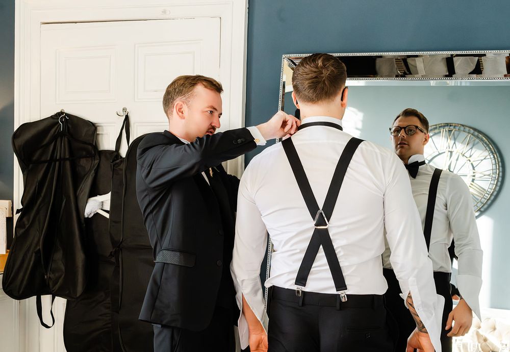 Groomsmen in formal attire help each other adjust suspenders and bowties while getting ready for a wedding ceremony.