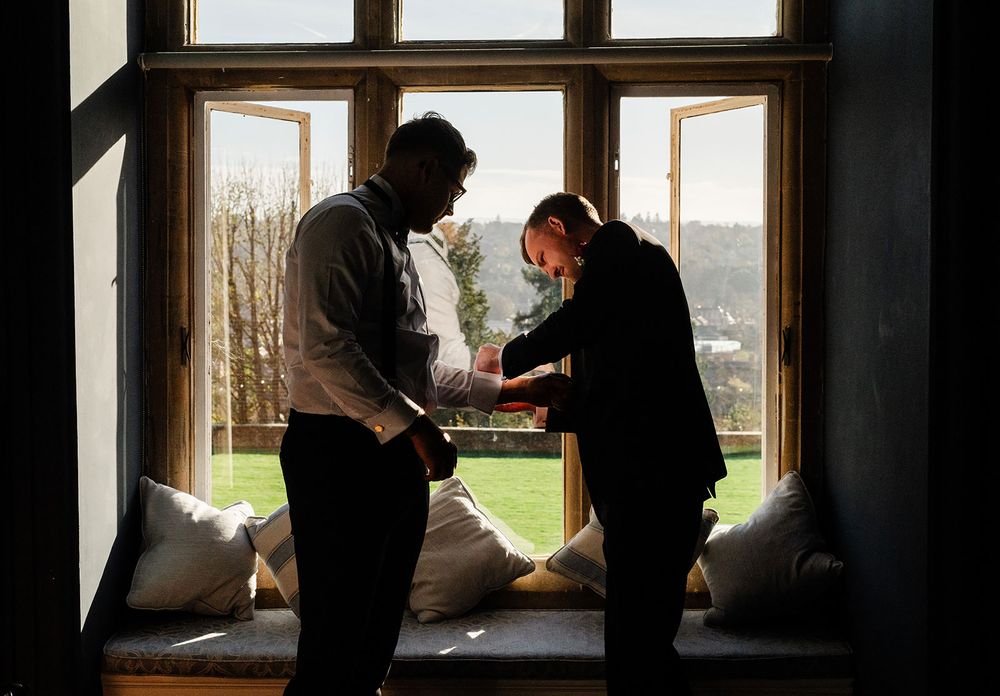 Two silhouetted figures stand by a window working together in a dimly lit room with natural light streaming in.