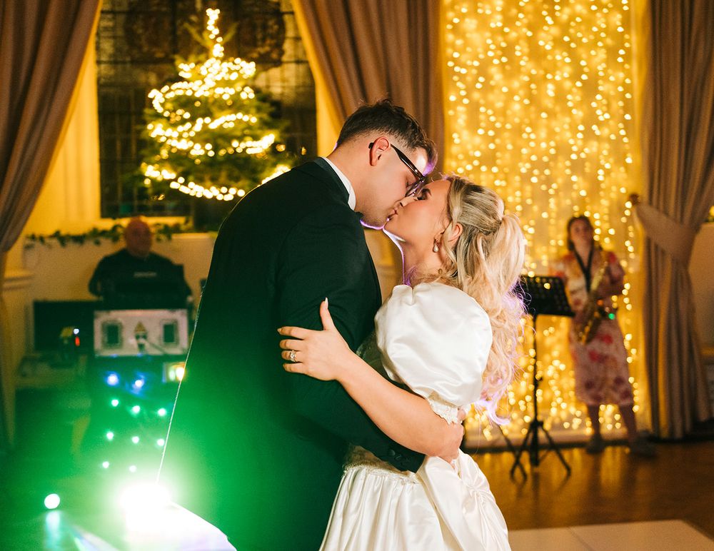 A couple shares a romantic dance at their wedding reception with twinkling lights and Christmas trees in the background.